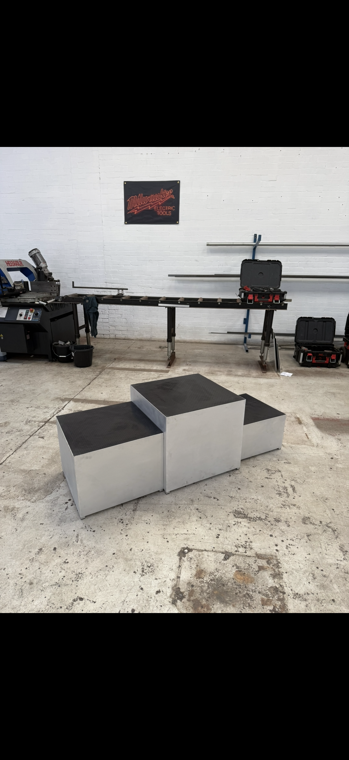 A metal podium with three steps on a concrete floor inside a workshop, with a white brick wall and tools, including a Milwaukee sign, in the background.