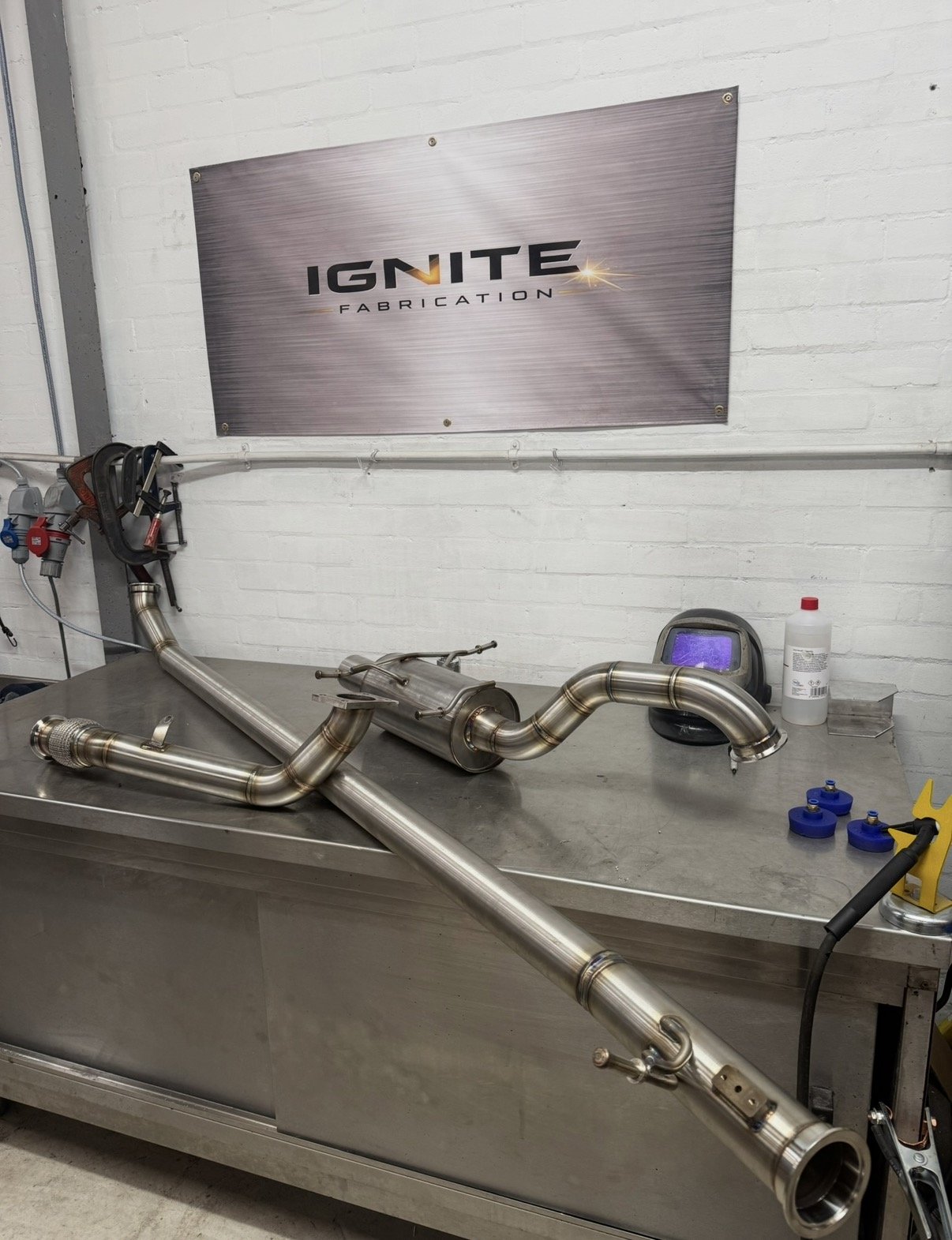 Metal automotive exhaust pipe on worktable in a workshop with a white brick wall and a sign reading 'IGNITE FABRICATION' in the background.