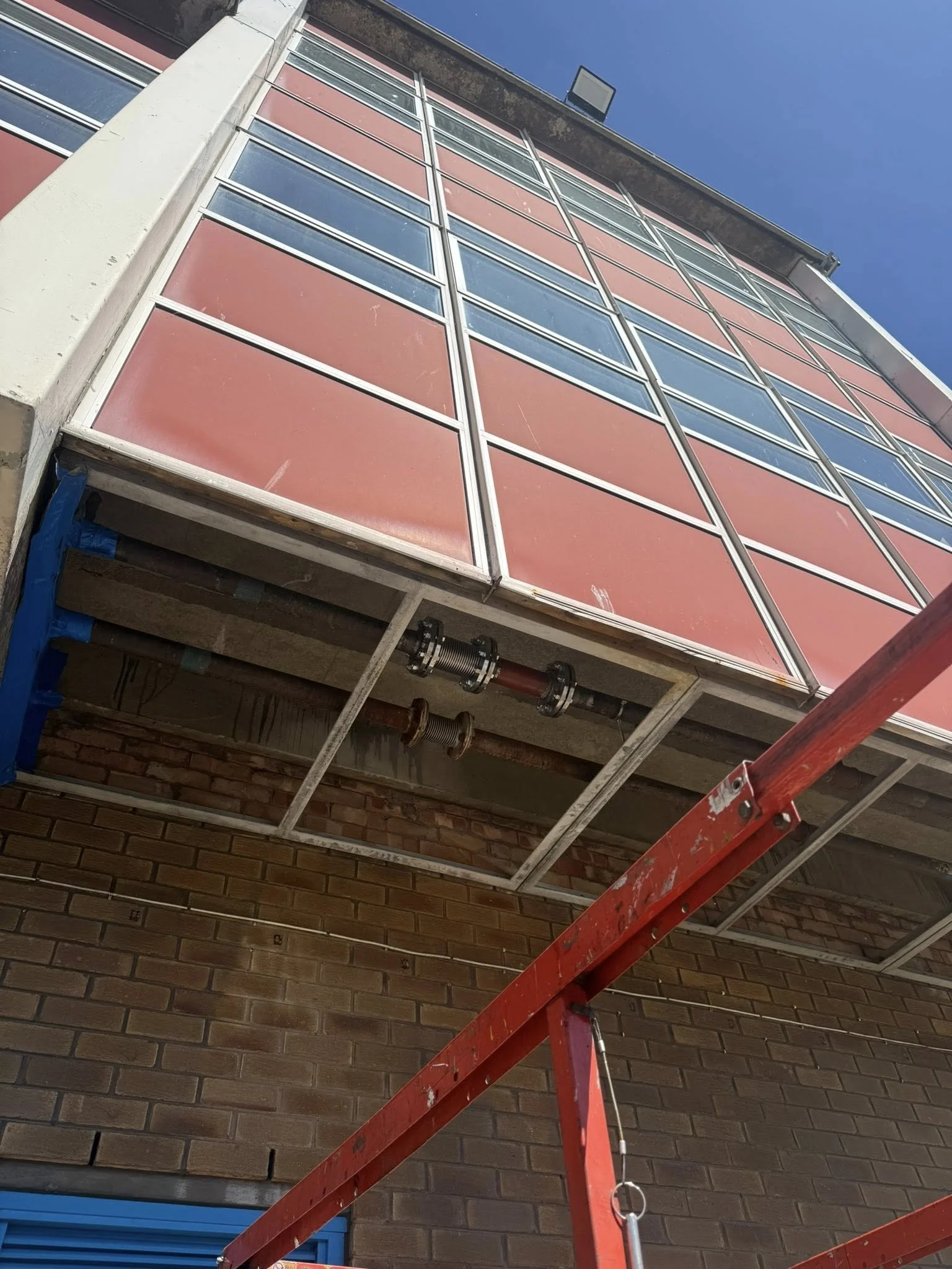 The image shows a side view of a multi-story building with a brick exterior, large windows, and a metal framework that appears to be under construction or renovation. There is scaffolding or a ladder with a red base in the foreground, and the sky is clear and blue.