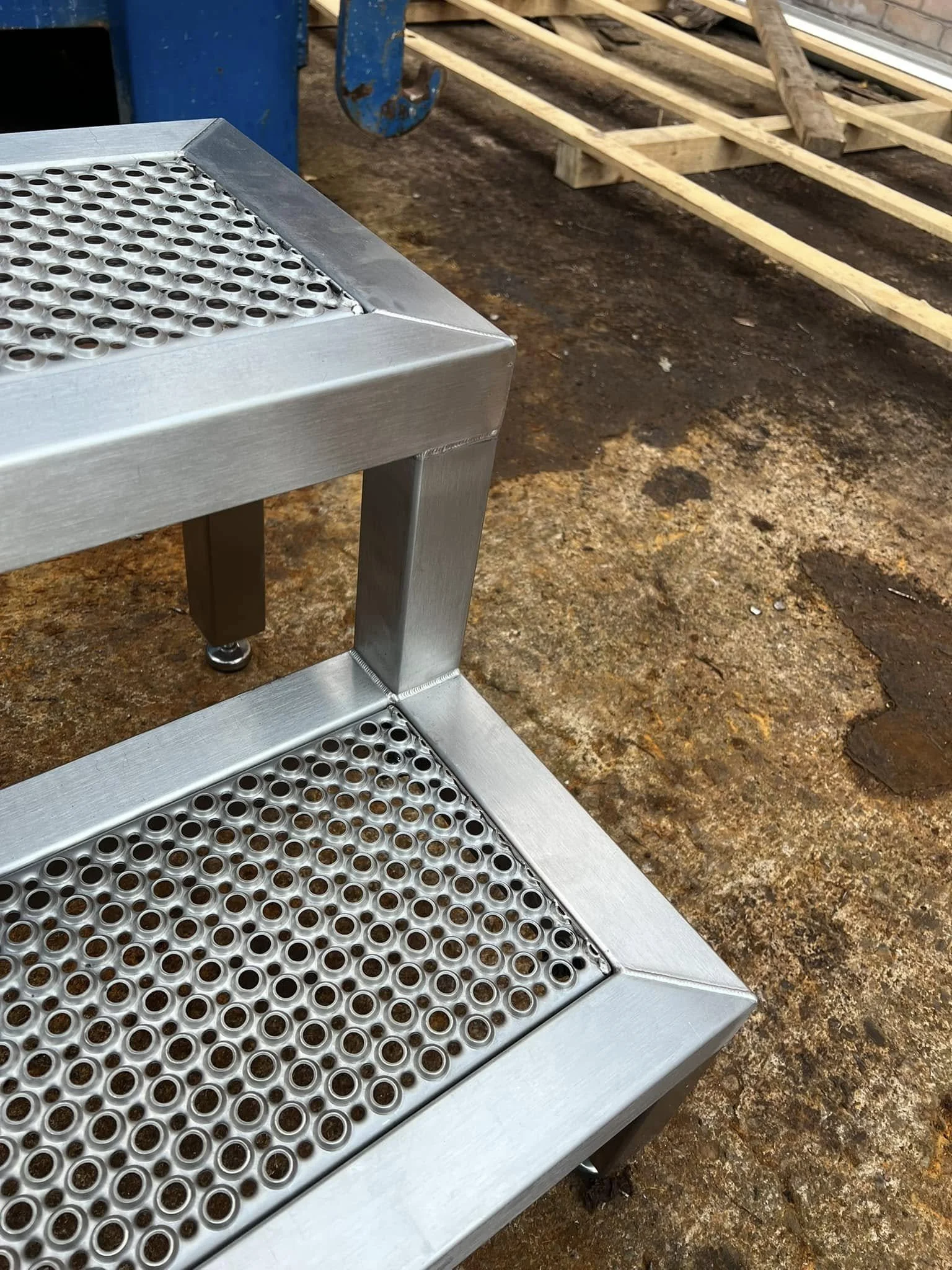 A metal workbench with mesh surface in a workshop, with a wooden frame and bricks in the background.