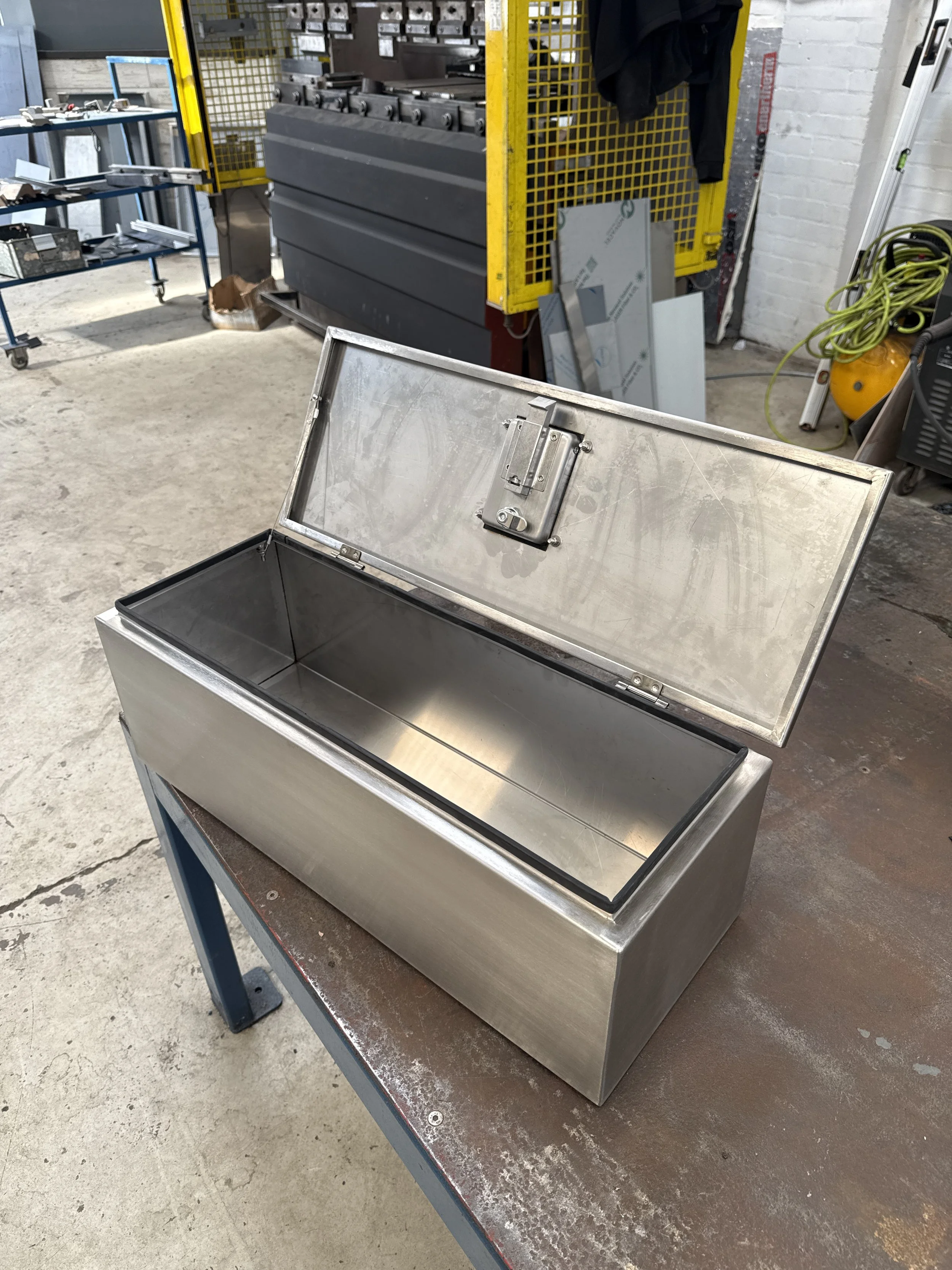 An open stainless steel box with a hinged lid, situated on a dusty workbench in a workshop or industrial space.