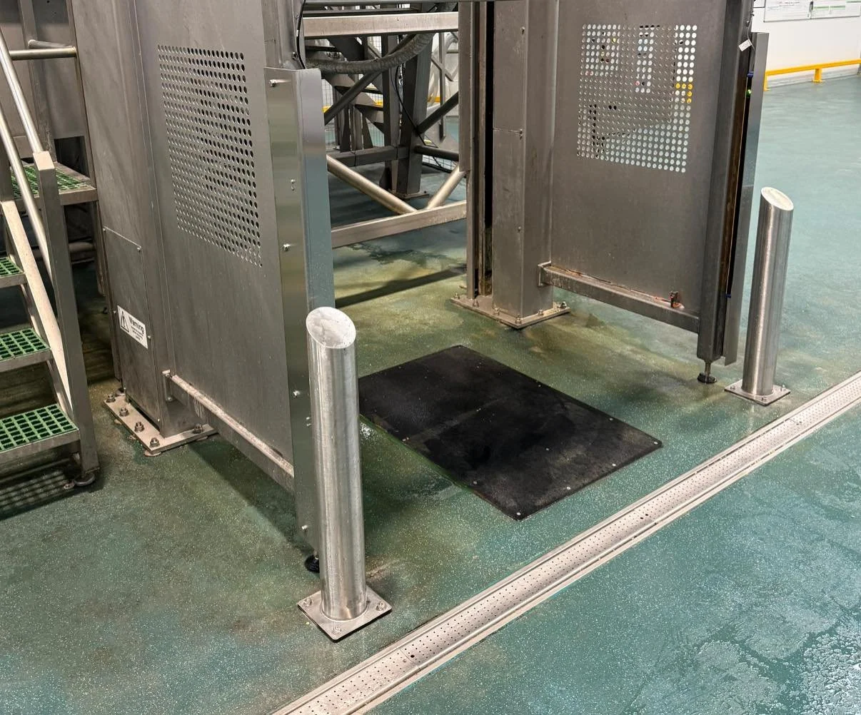 Industrial equipment with a black mat on the floor, surrounded by metal posts in a facility with a green speckled floor and a yellow safety barrier in the background.