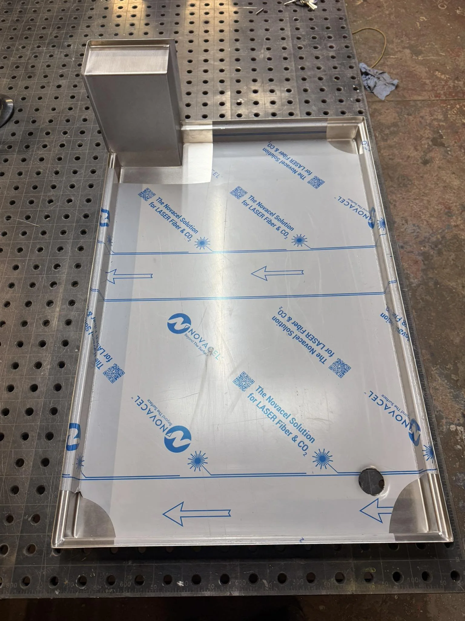 A metal tray with blue text and arrows, placed on a black perforated work surface. The tray has a raised edge and a small rectangular block attached to one side.