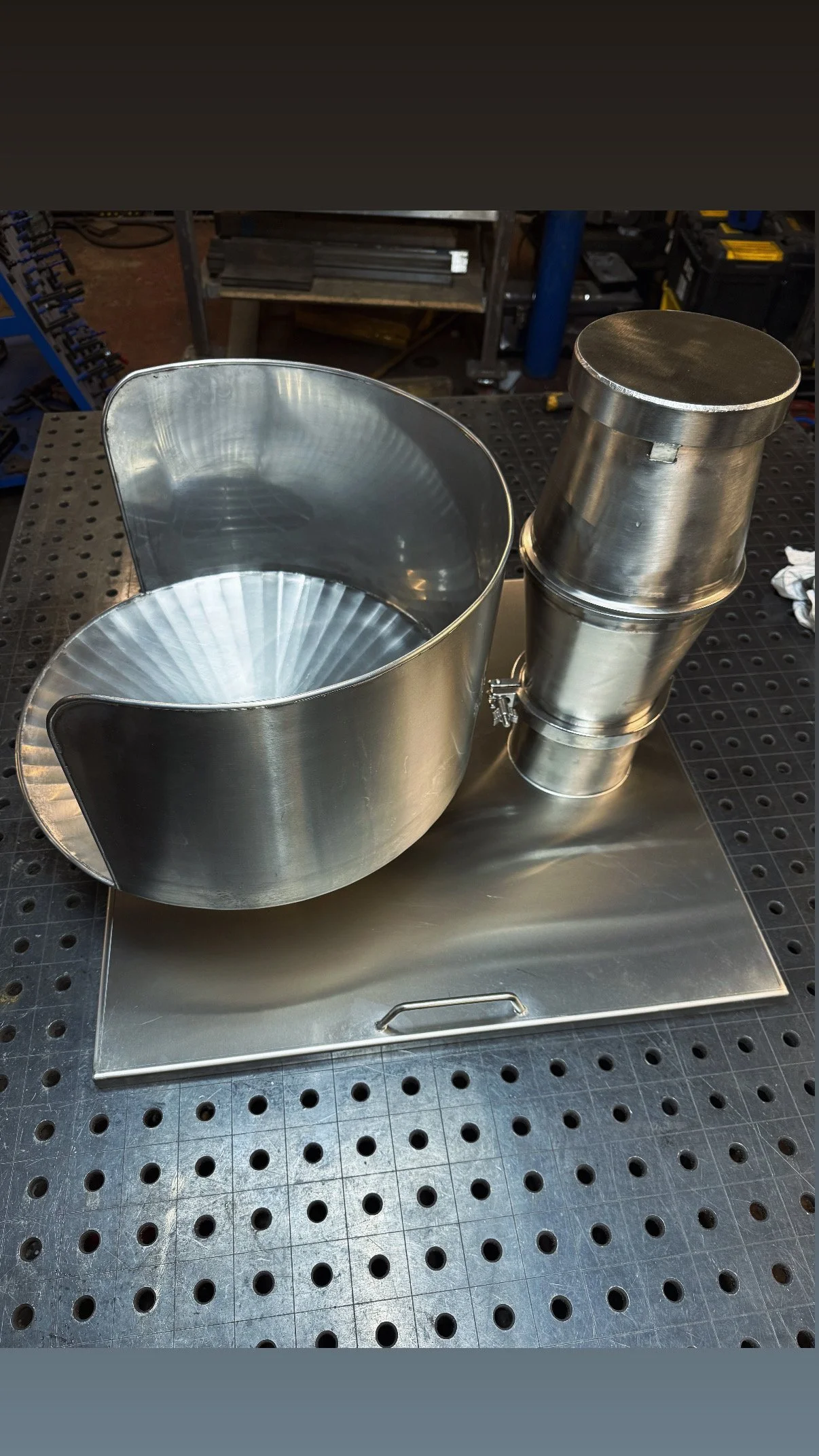 A stainless steel industrial piece of equipment, possibly a centrifuge or a similar device, with a large round basin and a cylindrical component attached to the side, on a perforated metal work surface in a workshop or manufacturing environment.
