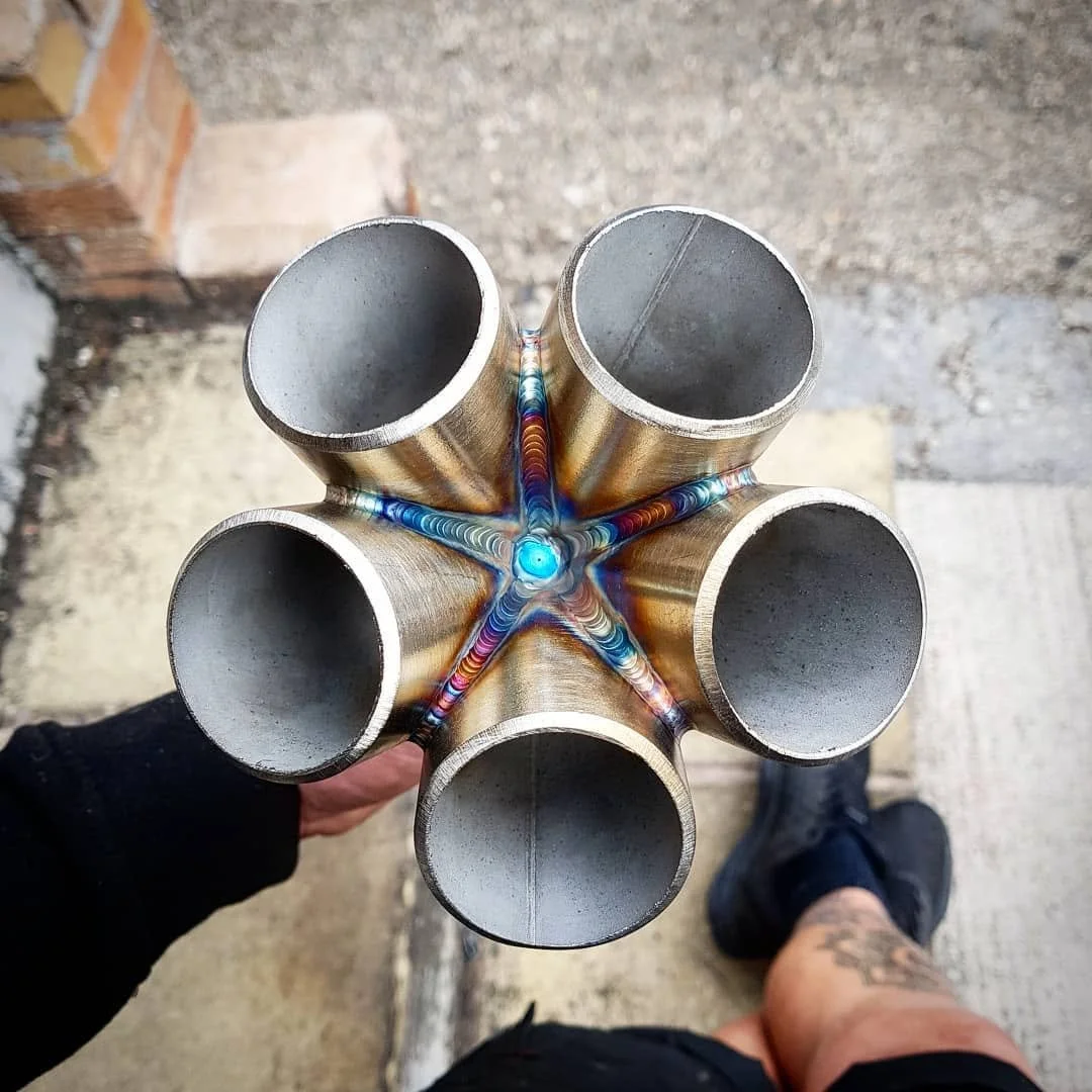 A person holding a metallic star-shaped pipe with six cylindrical openings, showing colorful welding at the joints, against a rough outdoor surface background.