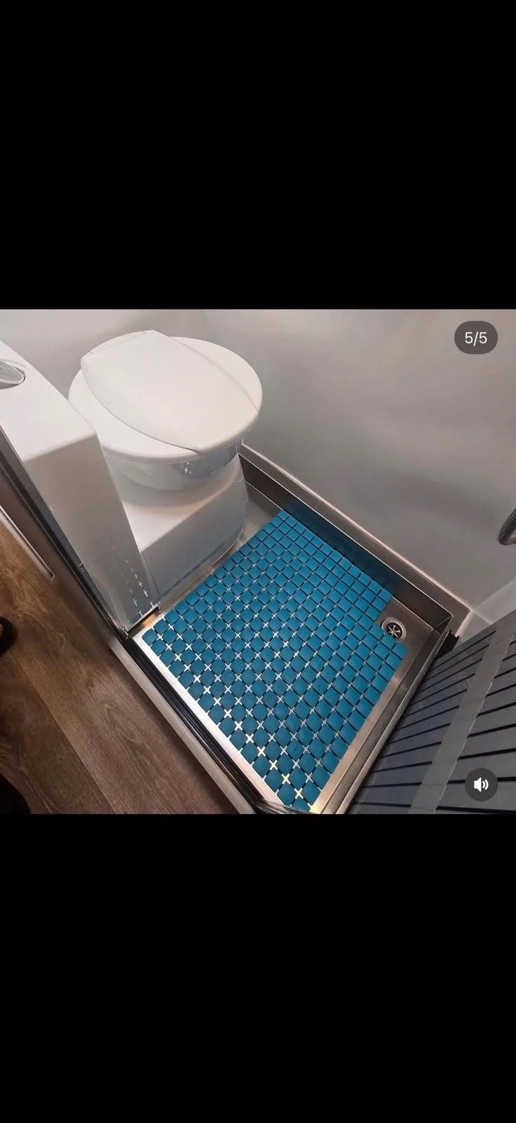 A compact bathroom with a white toilet, blue rubber mat inside a small shower area, and wooden flooring outside the shower.