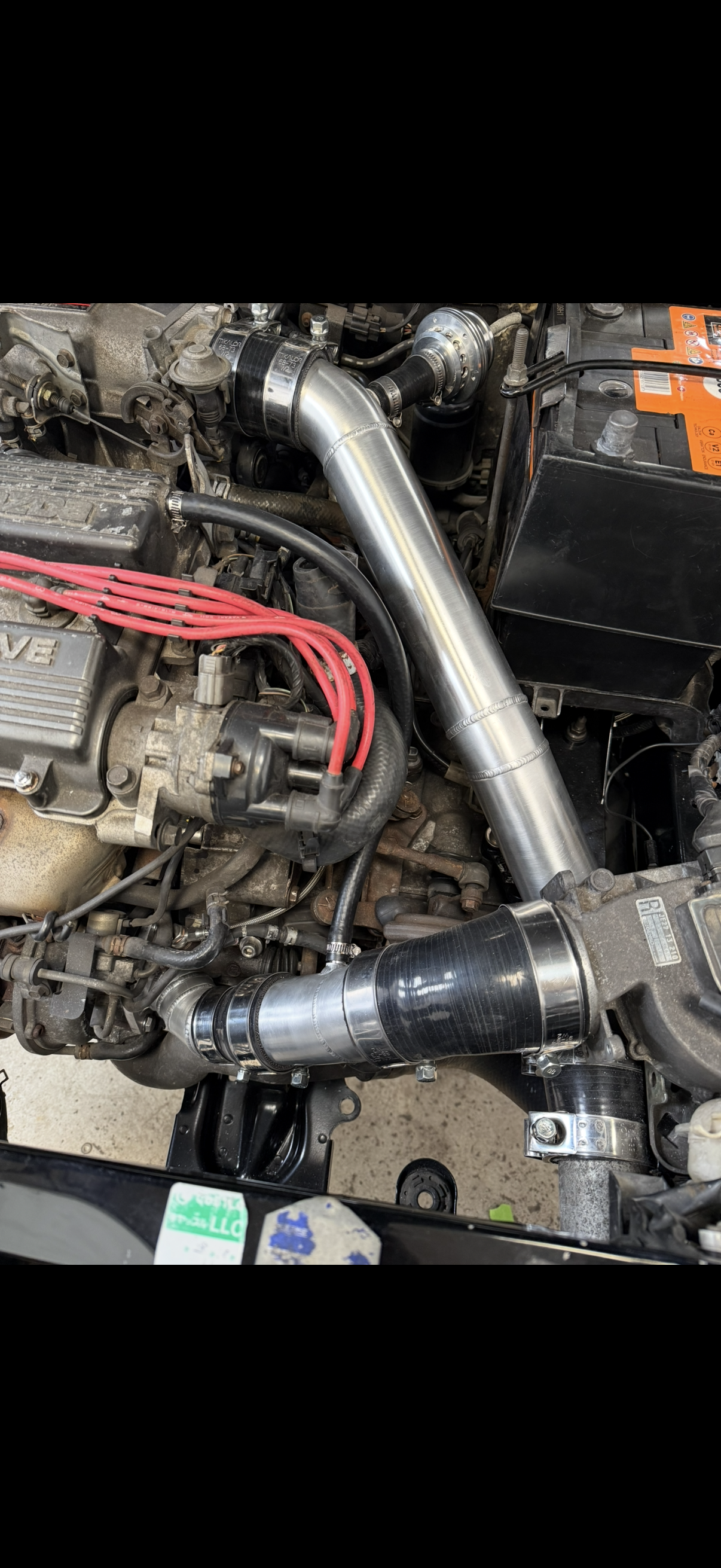 Inside a car engine bay showing a large turbocharger connected to the intake with shiny metal piping, a battery, and various engine components and wiring.