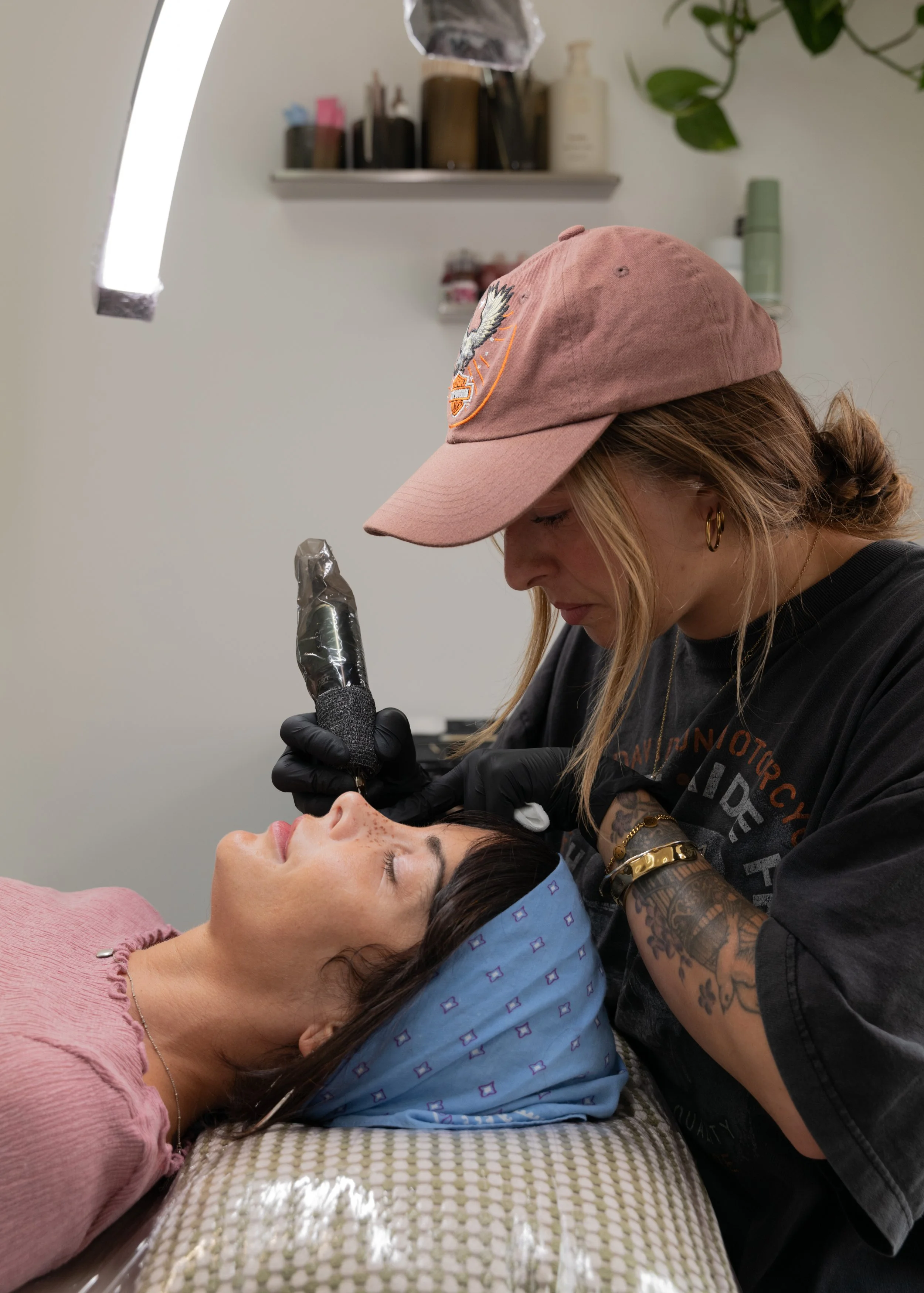 A woman lying down with a blue headscarf receives a faux freckle tattoos from a tattoo artist who is wearing a pink cap and black gloves in a tattoo studio.