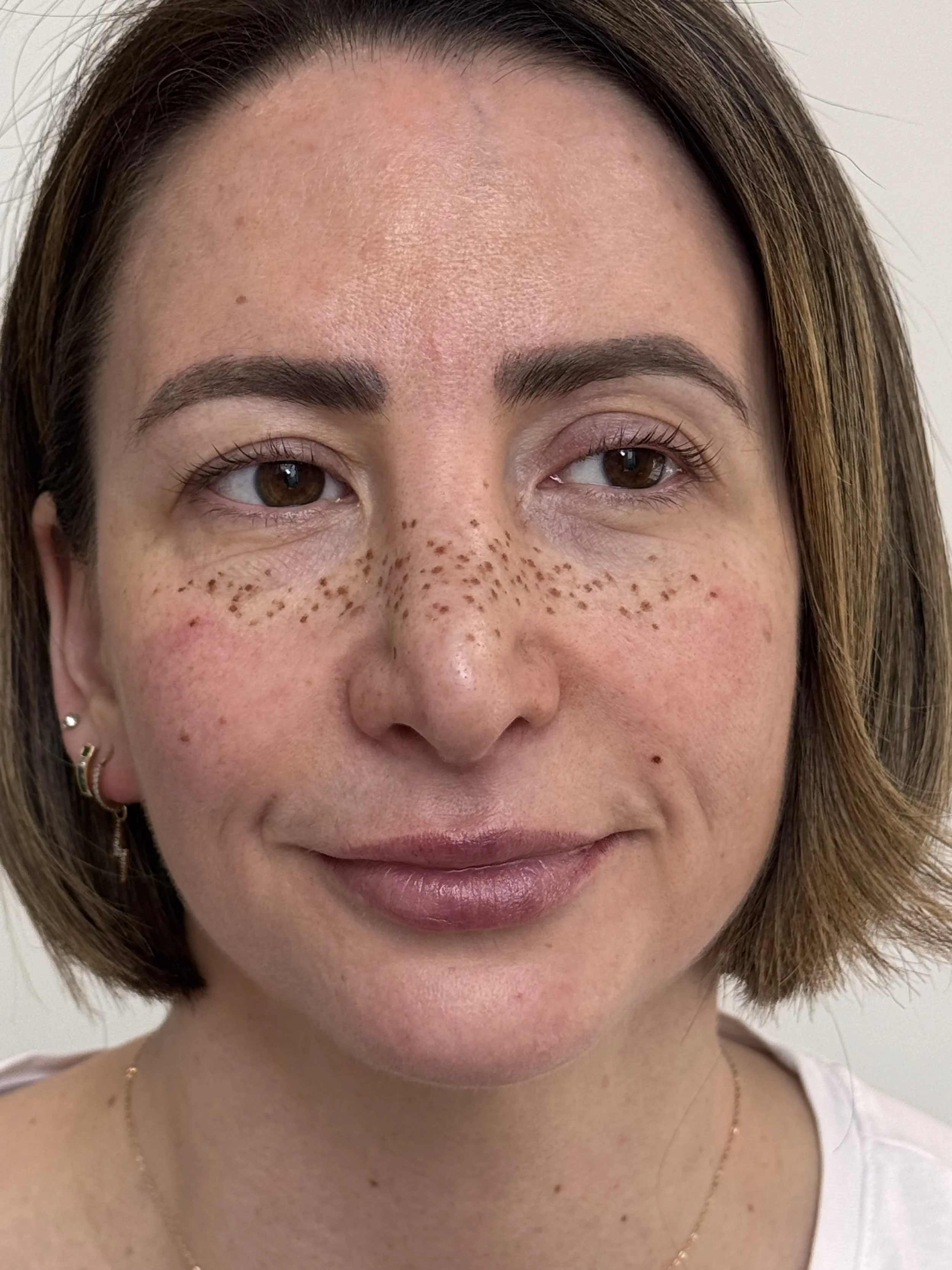 A woman with short brown hair, freckles across her nose and cheeks, wearing a white shirt and a delicate gold necklace, standing against a plain white background.