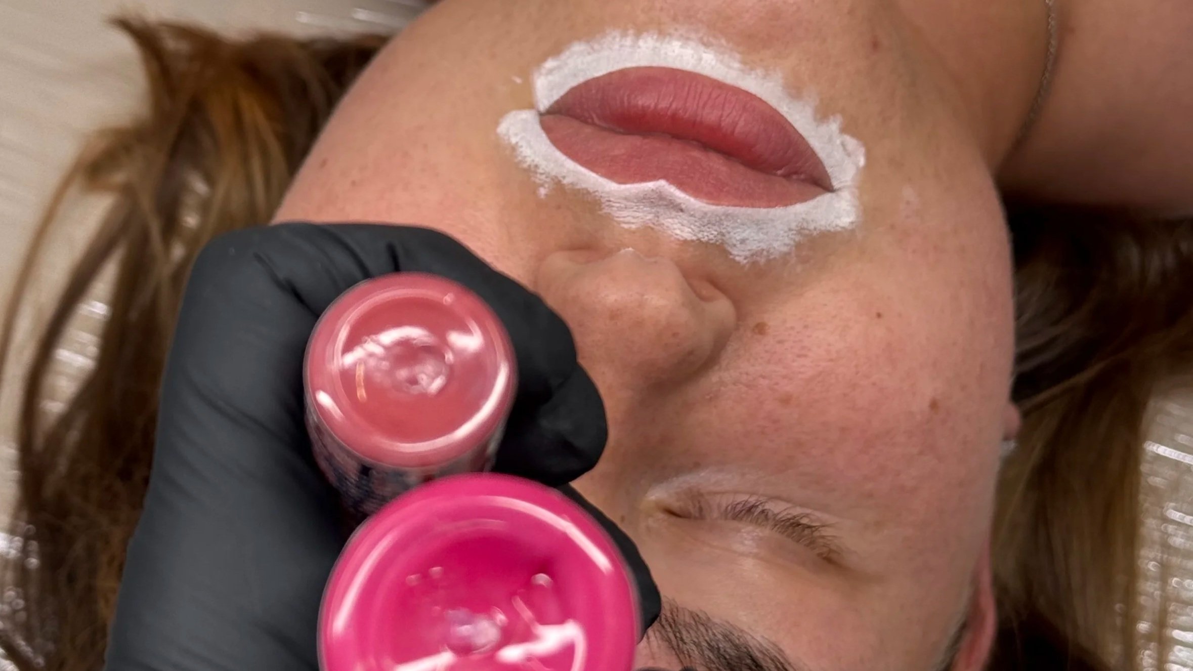 A woman lying down getting her lips and around her mouth painted with a pink lipstick by a person wearing black gloves.