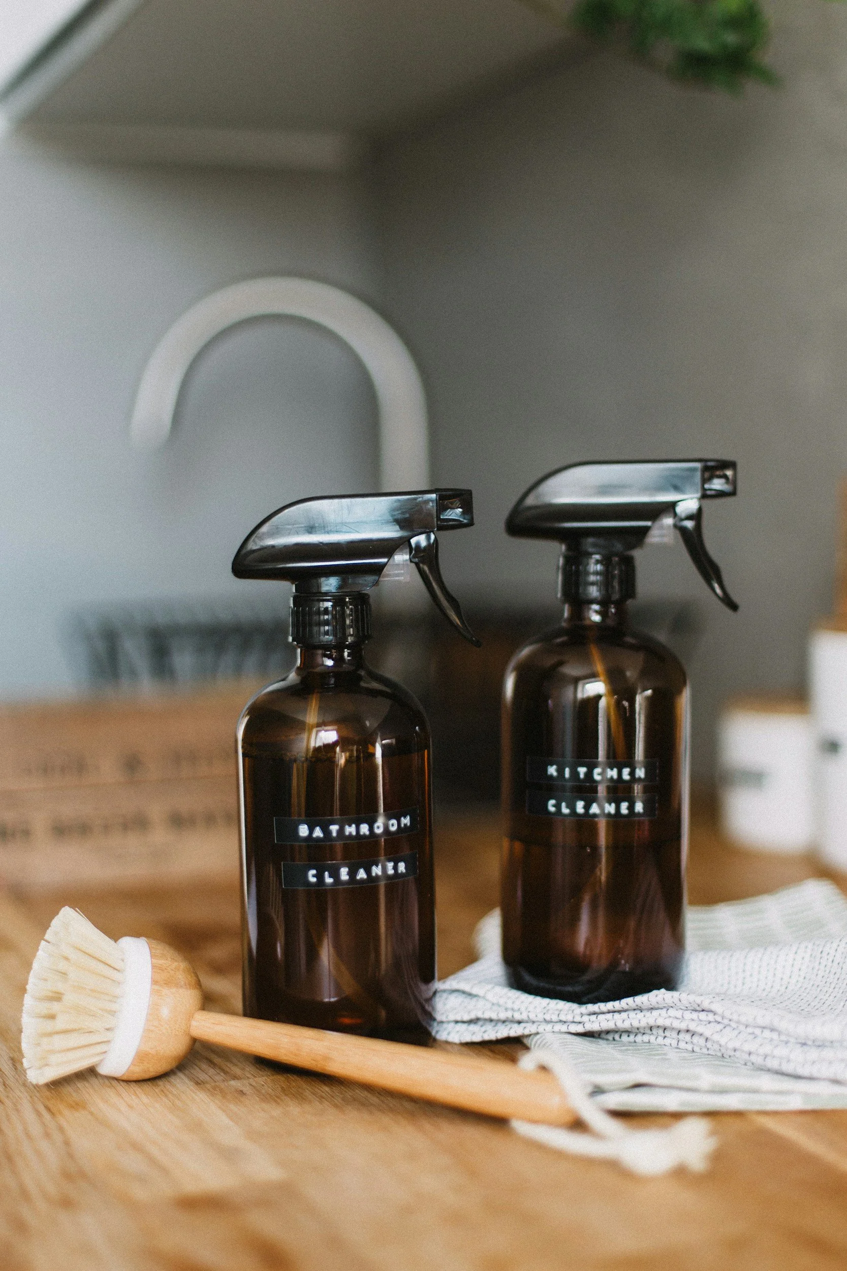Two brown bottles labeled 'Bathroom Cleaner' and 'Kitchen Cleaner' with black caps, placed on a wooden surface with a white towel underneath, and a natural bristle brush with a wooden handle in front of them.