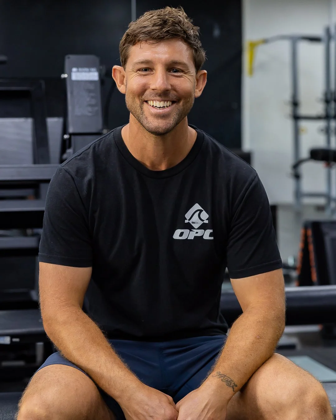 A smiling man with short brown hair sitting in a gym, wearing a black OPC t-shirt and dark shorts, with gym equipment in the background.