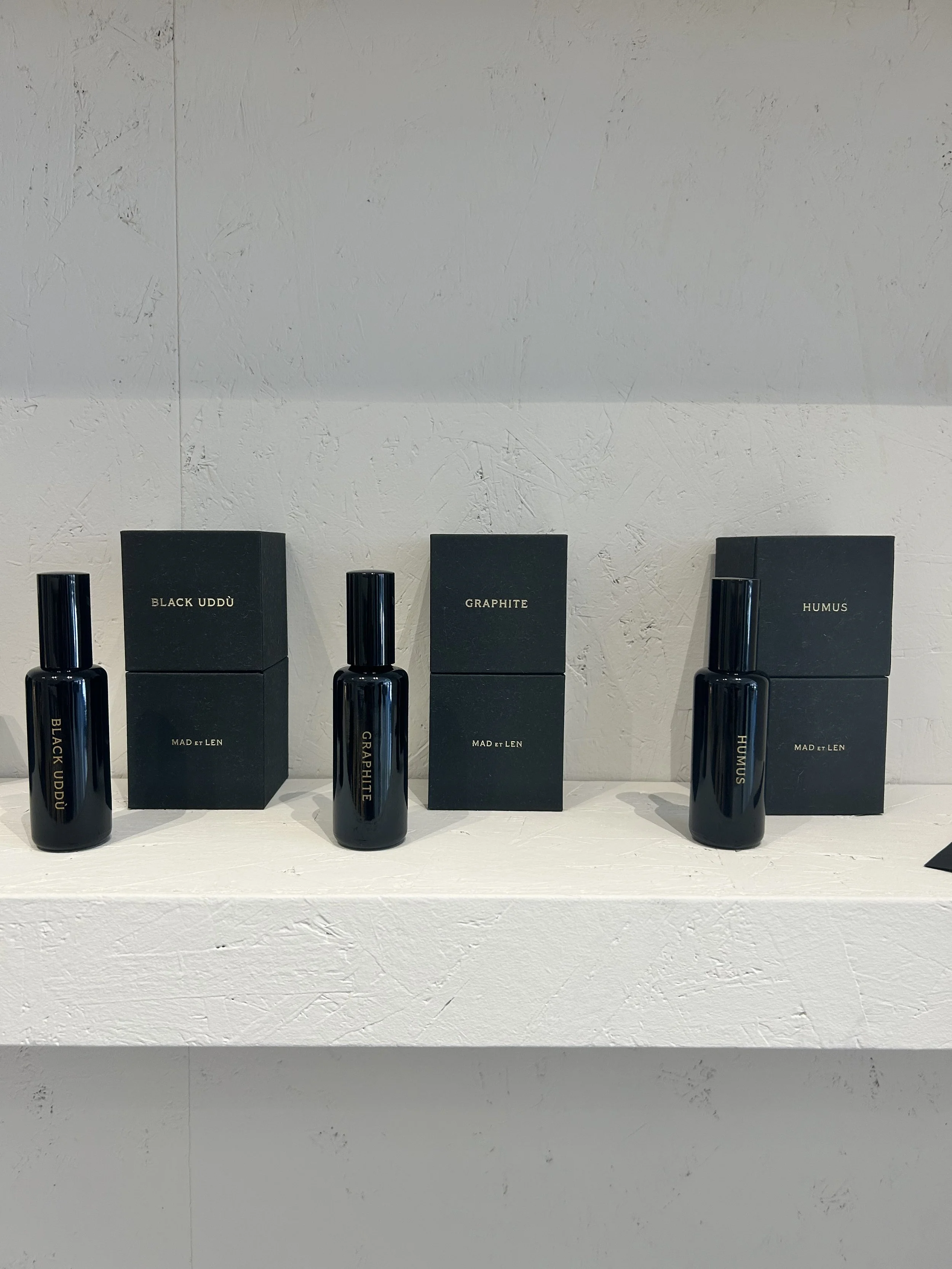 Cuatro frascos de perfume con cajas negras y etiquetas: Black Uddù, Graphite, Humus y Mad et Len, en una mesa blanca con pared de fondo de color gris claro.