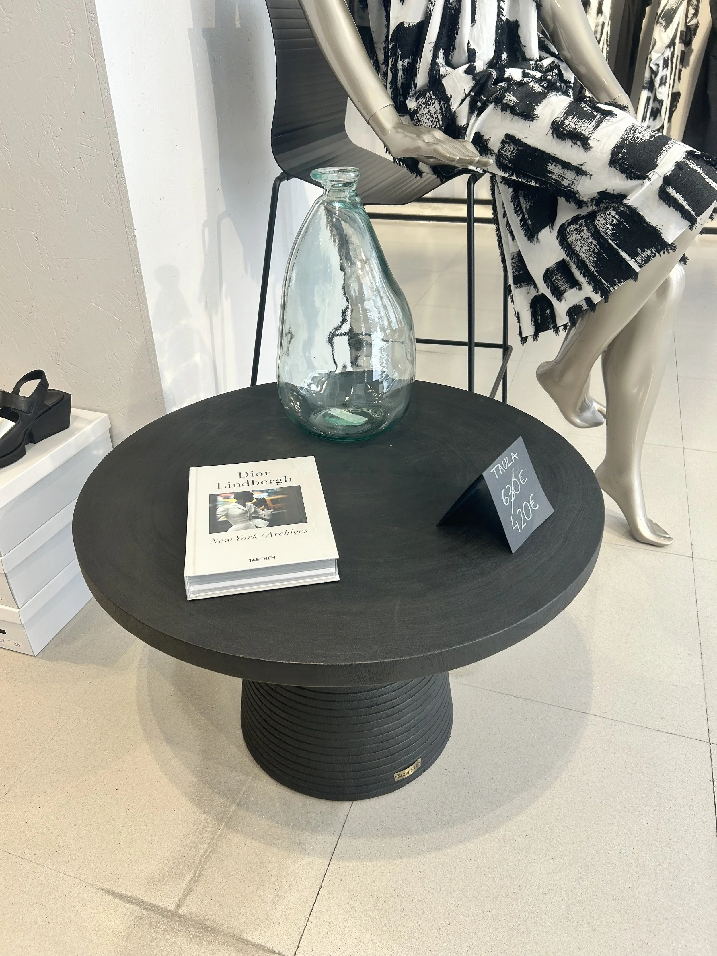 Una mesa redonda negra con un libro, un cartel con precios y un jarrón de vidrio en la superficie. En el fondo, una figura de maniquí con ropa blanca y negra, y una silla negra con patas de metal.
