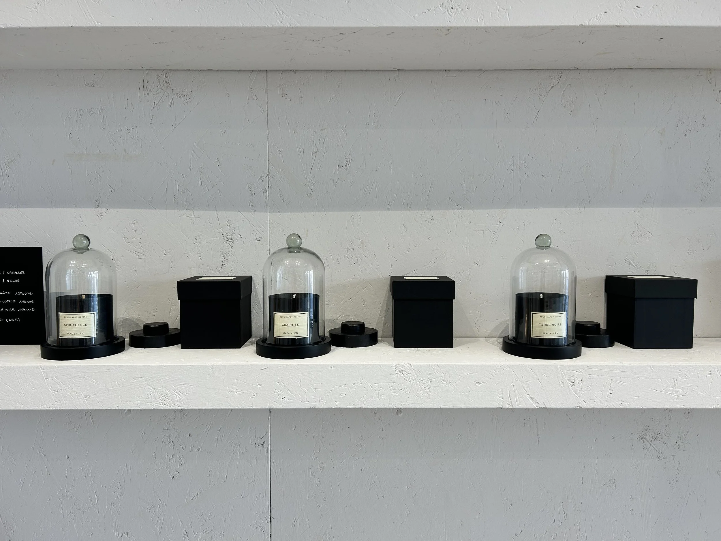 Muestra una estantería blanca con varios frascos de cristal con tapas negras, etiquetados con diferentes nombres de minerales o perfumes, y tres cajas negras. La pared de fondo es blanca con textura.