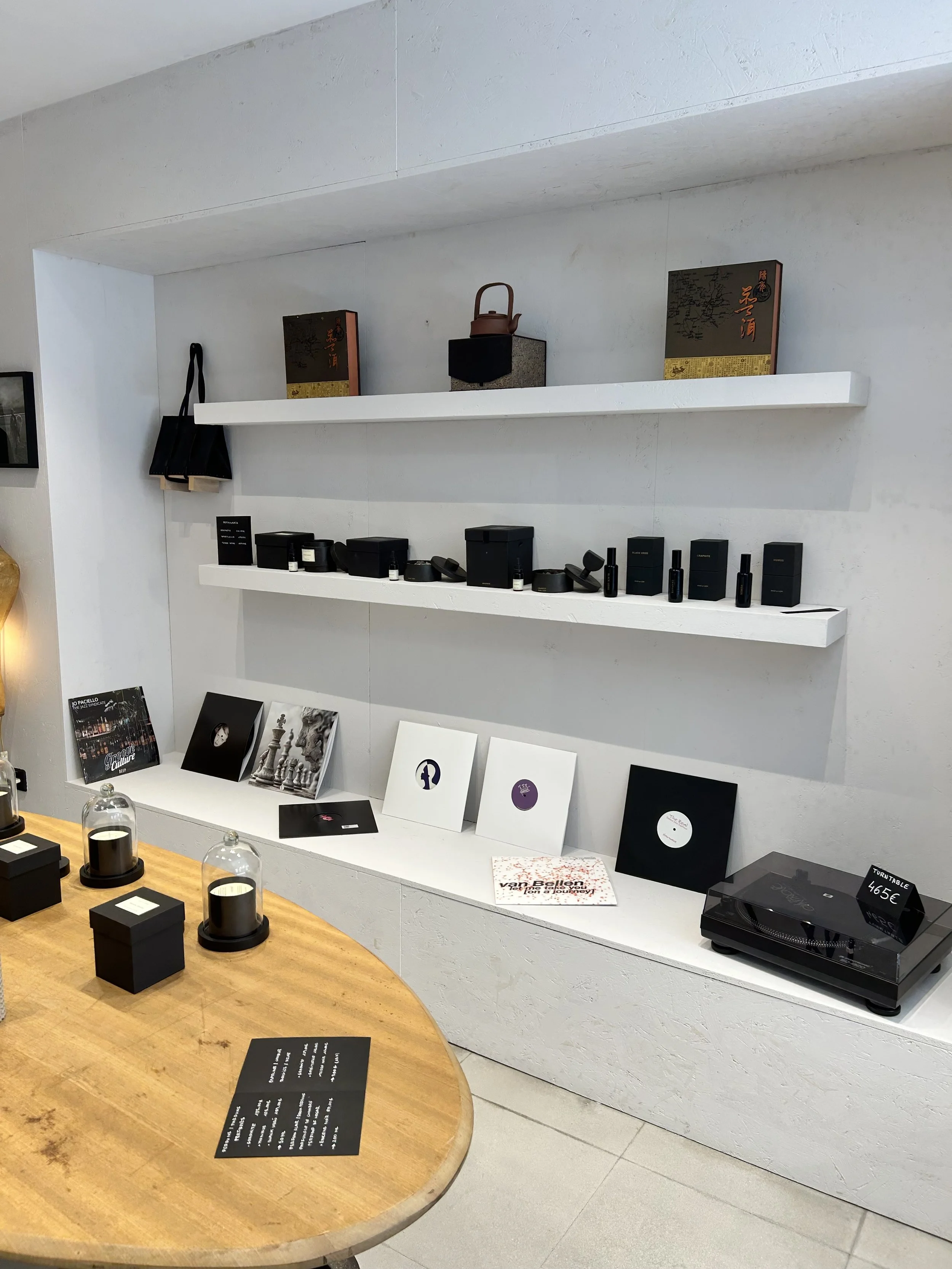 Imagen de una tienda con exhibición de productos de perfumes y fragancias en estanterías blancas. Hay una mesa de madera con velas negras y cartel con información, y en la pared hay dos estantes con cajas decorativas y un tetera en el centro.