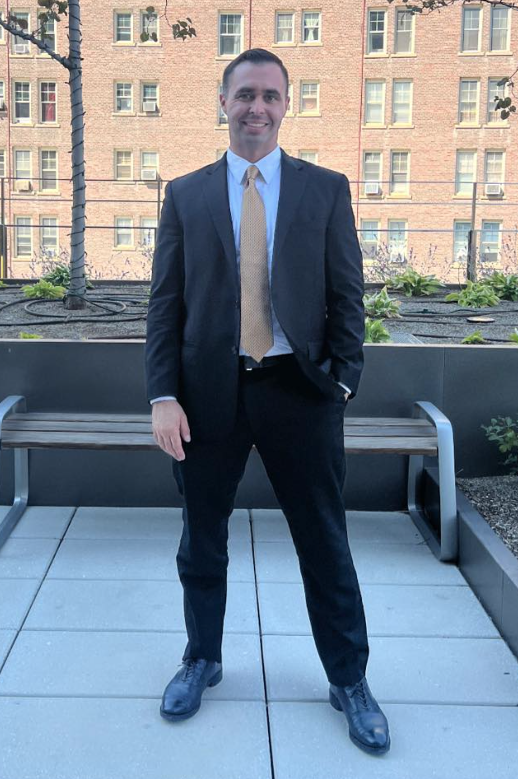 A man in a black suit, white shirt, and beige tie standing outdoors on a balcony with modern buildings in the background.