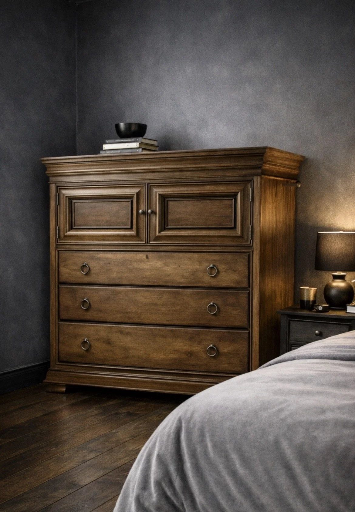 A wooden dresser with four drawers and a cabinet on top, situated beside a nightstand with a lamp and decorative items, in a bedroom with dark walls.