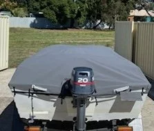 A boat covered with a gray tarp on a trailer in a grassy outdoor area.