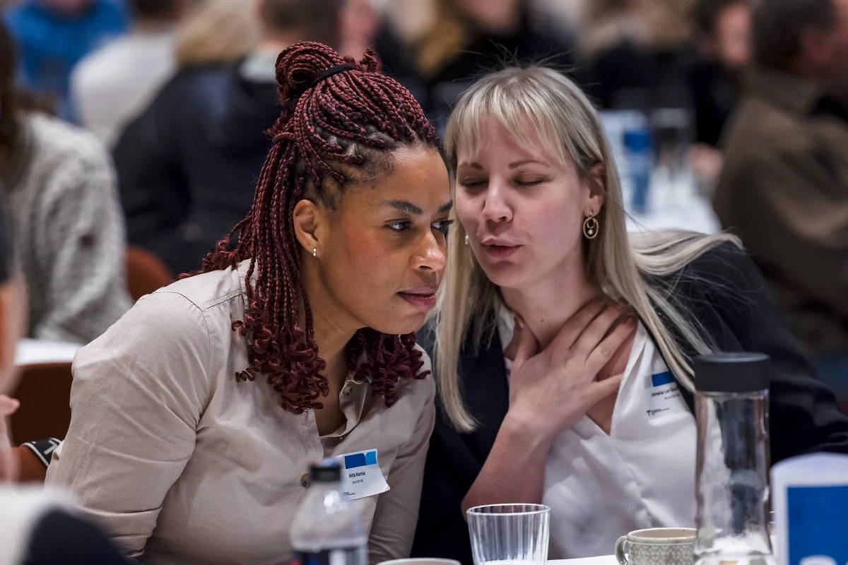 Two women having a serious conversation at a table in a crowded setting, possibly a conference or meeting.