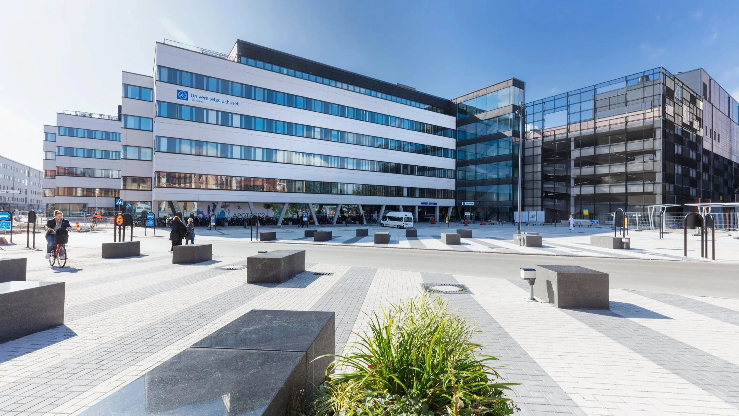 Modern hospital building with white and black glass facade, outdoor plaza with pedestrians, cyclist, and benches, clear blue sky.
