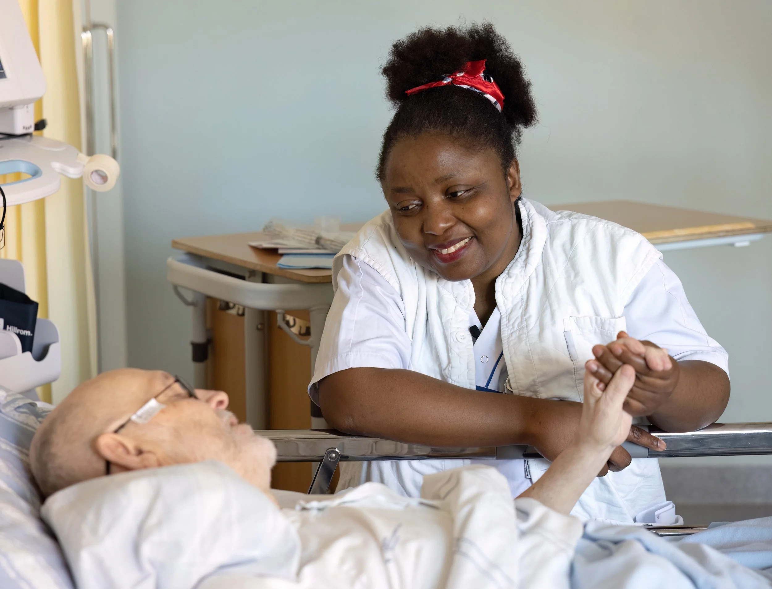 A healthcare worker with curly hair and a red bandana is smiling and arm-wrestling an elderly patient in a hospital room.