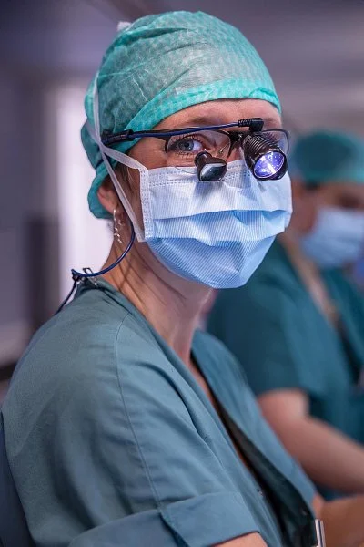 A female healthcare professional in surgical scrubs, wearing a surgical cap, face mask, and magnifying loupes, with another healthcare worker in the background.