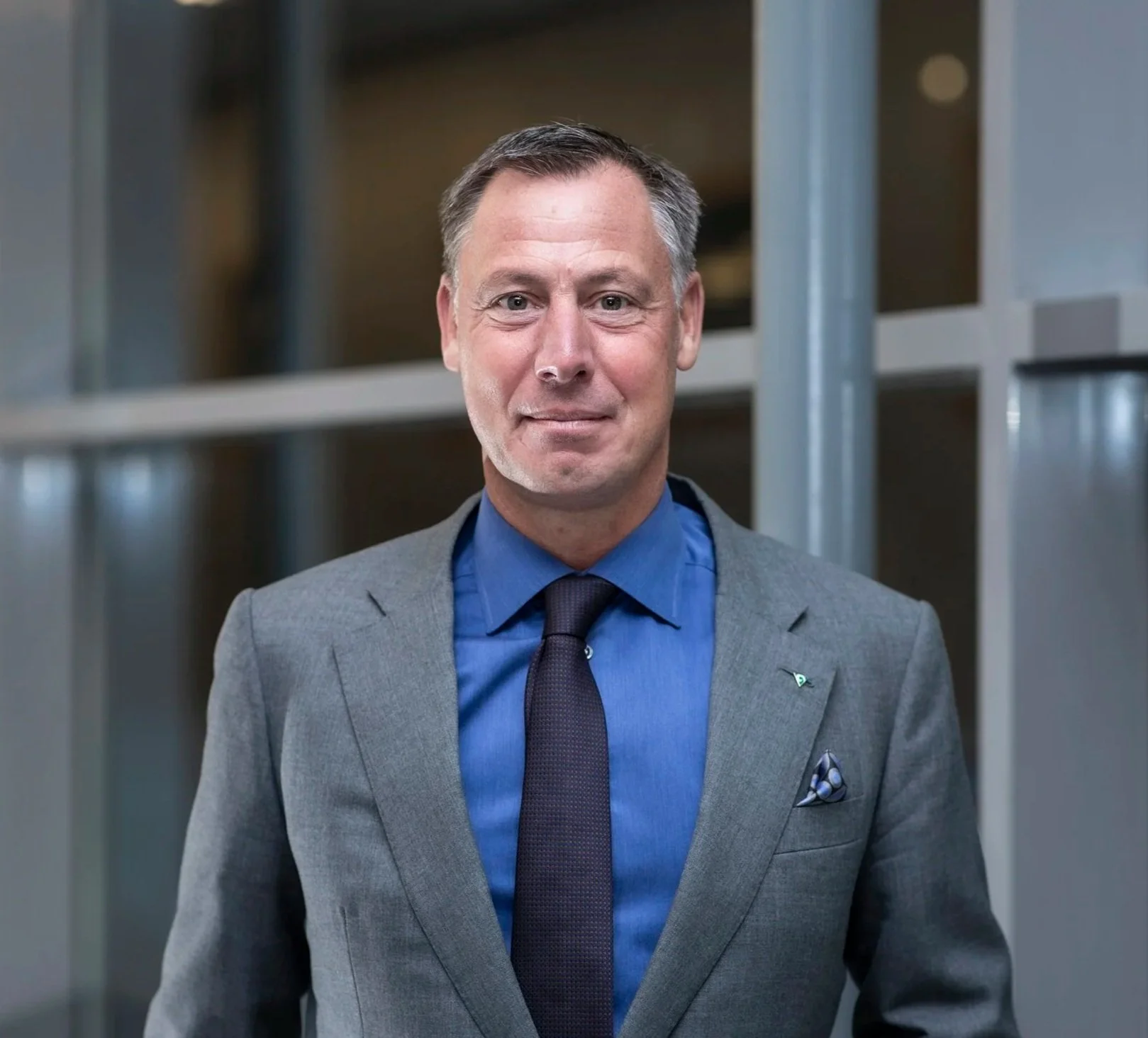 A man in a gray suit, blue shirt, and dark tie, standing in front of a modern office building with glass and metal panels, smiling at the camera.