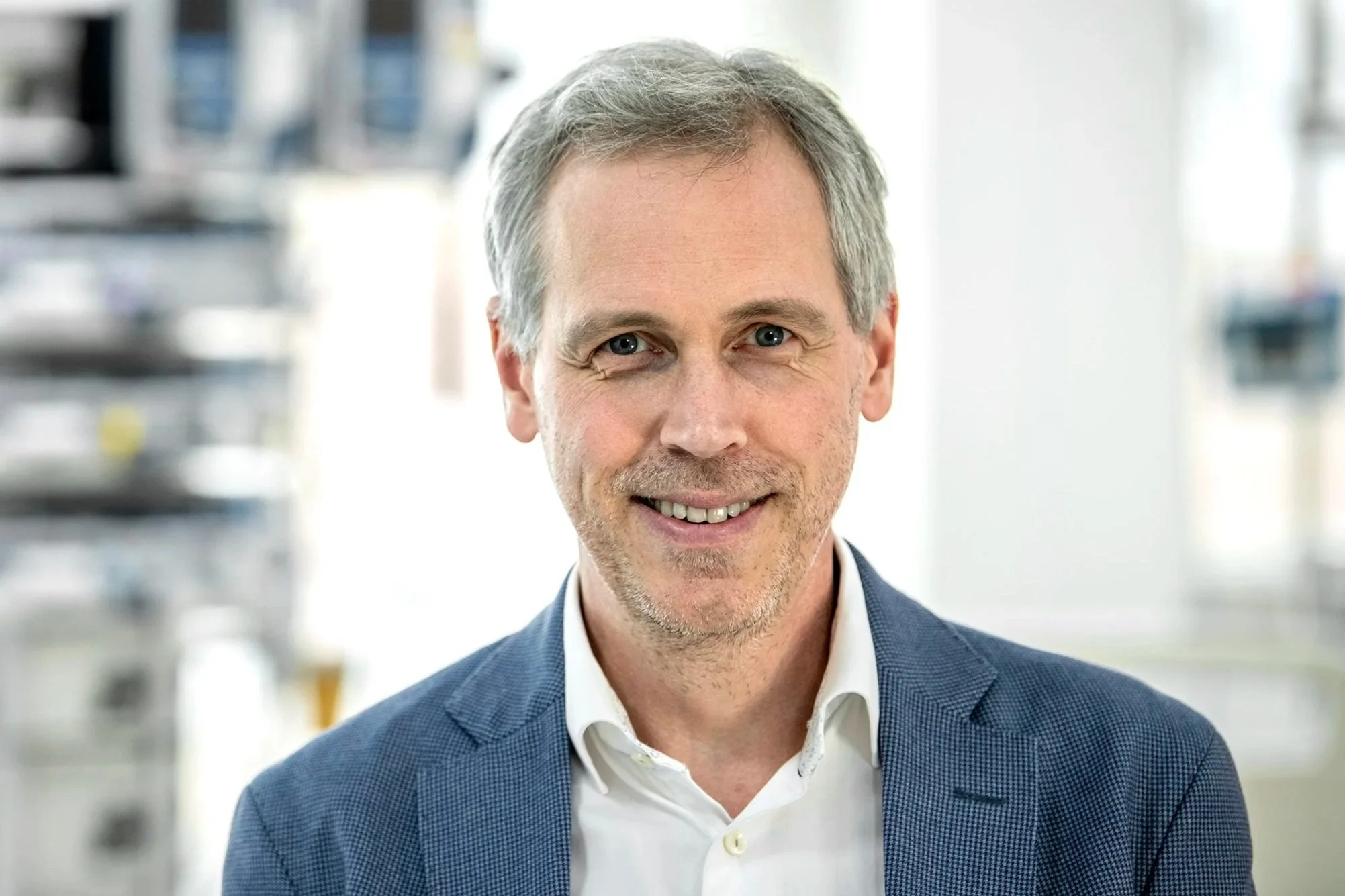 A middle-aged man with gray hair and blue eyes smiling, wearing a blue blazer over a white shirt, in a professional setting with shelves and folders in the background.