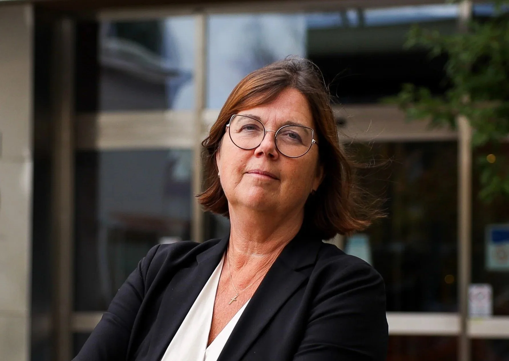 A middle-aged woman with glasses and shoulder-length brown hair standing outdoors wearing a black blazer over a white top, with a serious expression and her arms crossed.