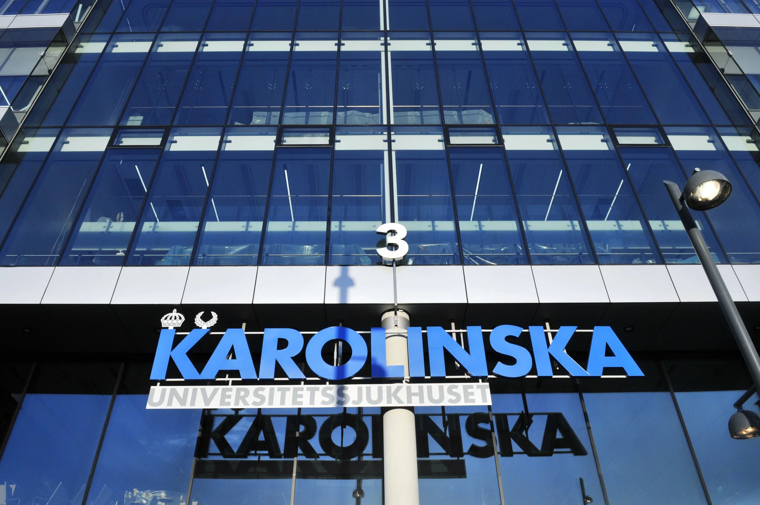 A modern glass building with the word 'KAROLINSKA' displayed prominently on a sign at the bottom, indicating a university hospital in Stockholm, Sweden.