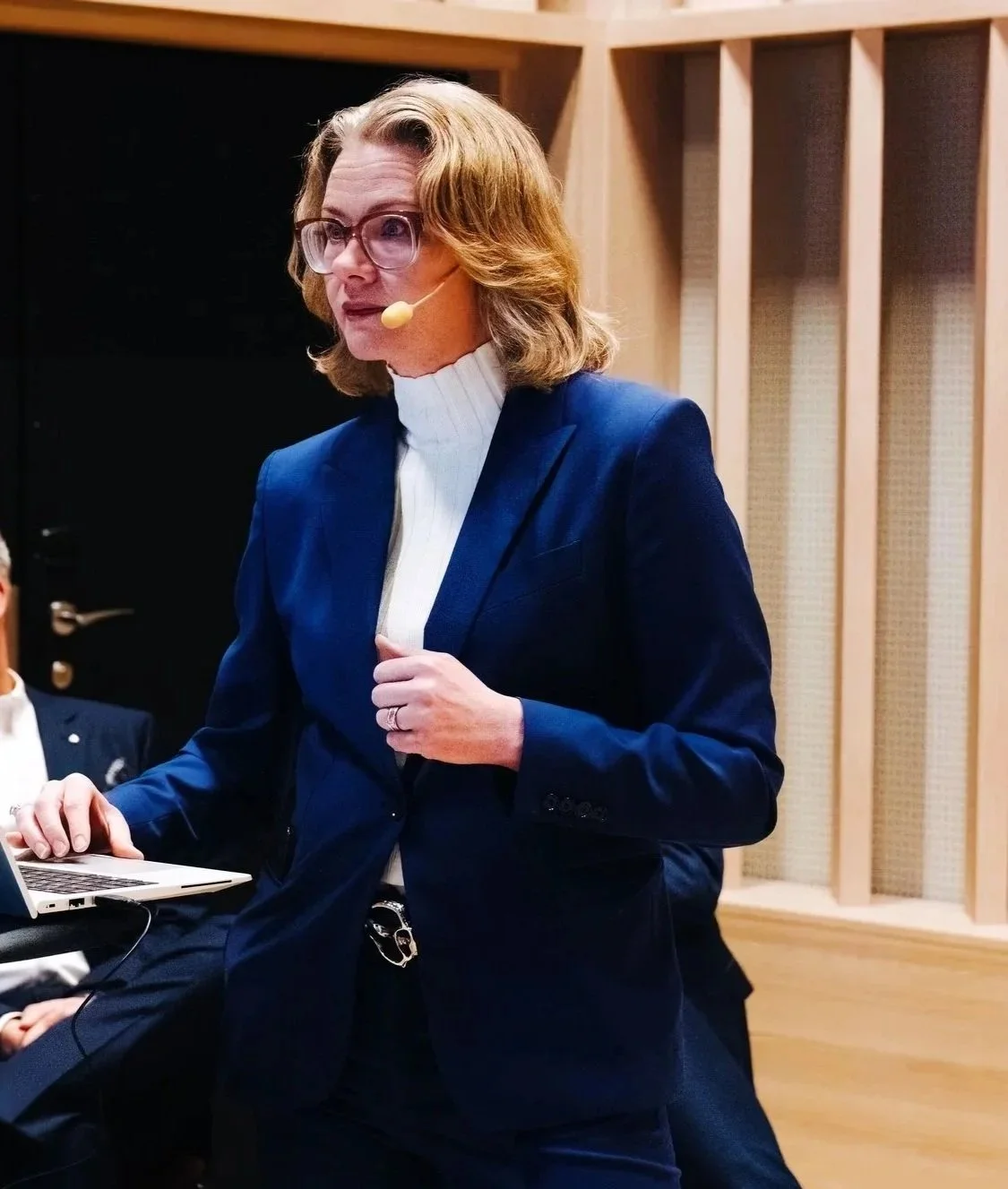 A woman in a blue suit with glasses and a headset standing in a recording studio or conference room.