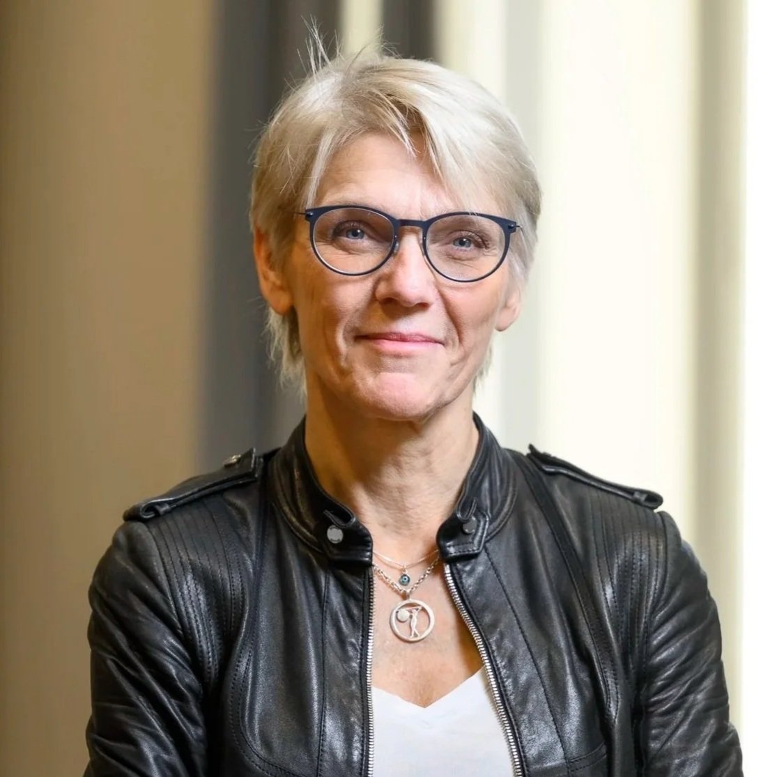 A middle-aged woman with short blonde hair, wearing glasses, a black leather jacket, and necklaces, standing indoors near a window.