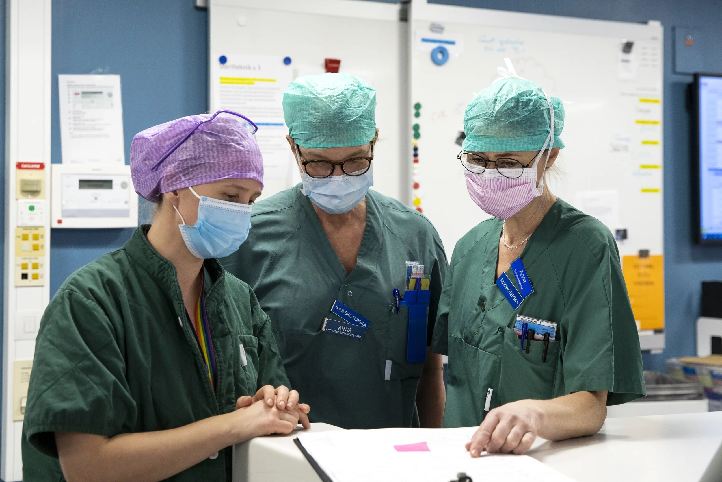Three healthcare professionals in scrubs, surgical masks, and caps gathered around and discussing medical notes or documents in a hospital setting.