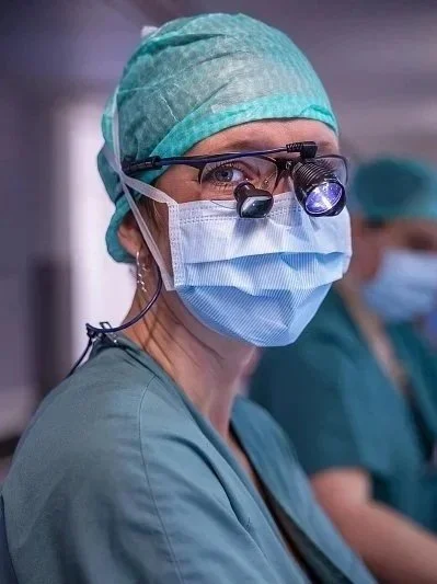 Close-up of a female surgeon in scrubs, wearing a surgical mask, head cap, and magnifying glasses with a light, in a sterile operating room.