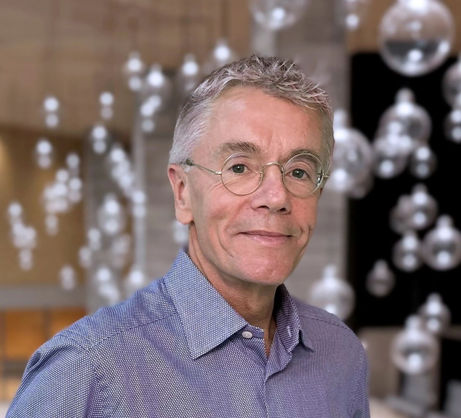 Portrait of an older man with gray hair and glasses, wearing a purple checkered shirt, standing in front of abstract art with metallic and wooden elements.