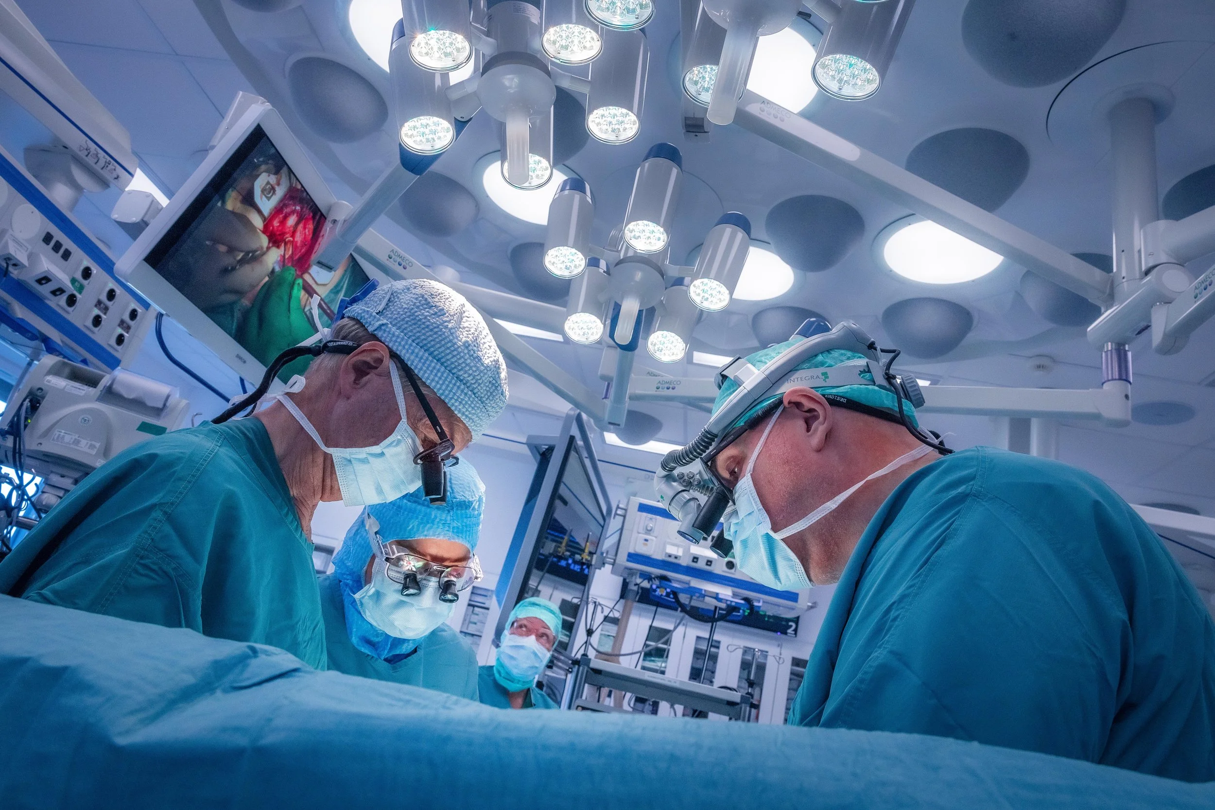 Surgeons performing surgery in an operating room under bright surgical lights, all wearing scrubs, masks, and protective gear.