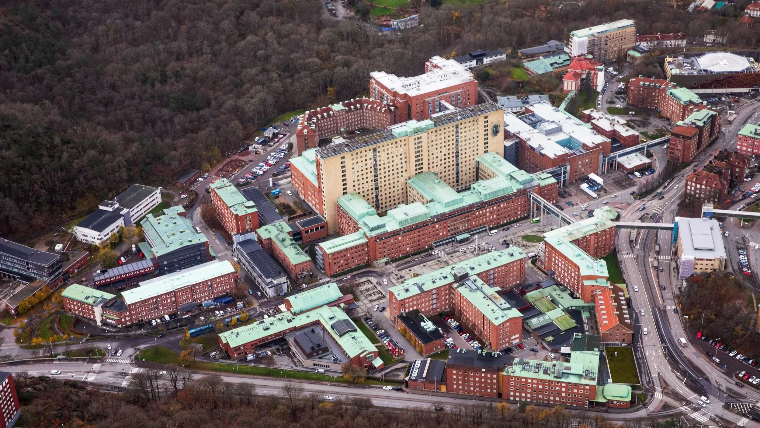Aerial view of a large hospital complex with multiple red-brick buildings and green rooftops, surrounded by roads, parking lots, and wooded areas.