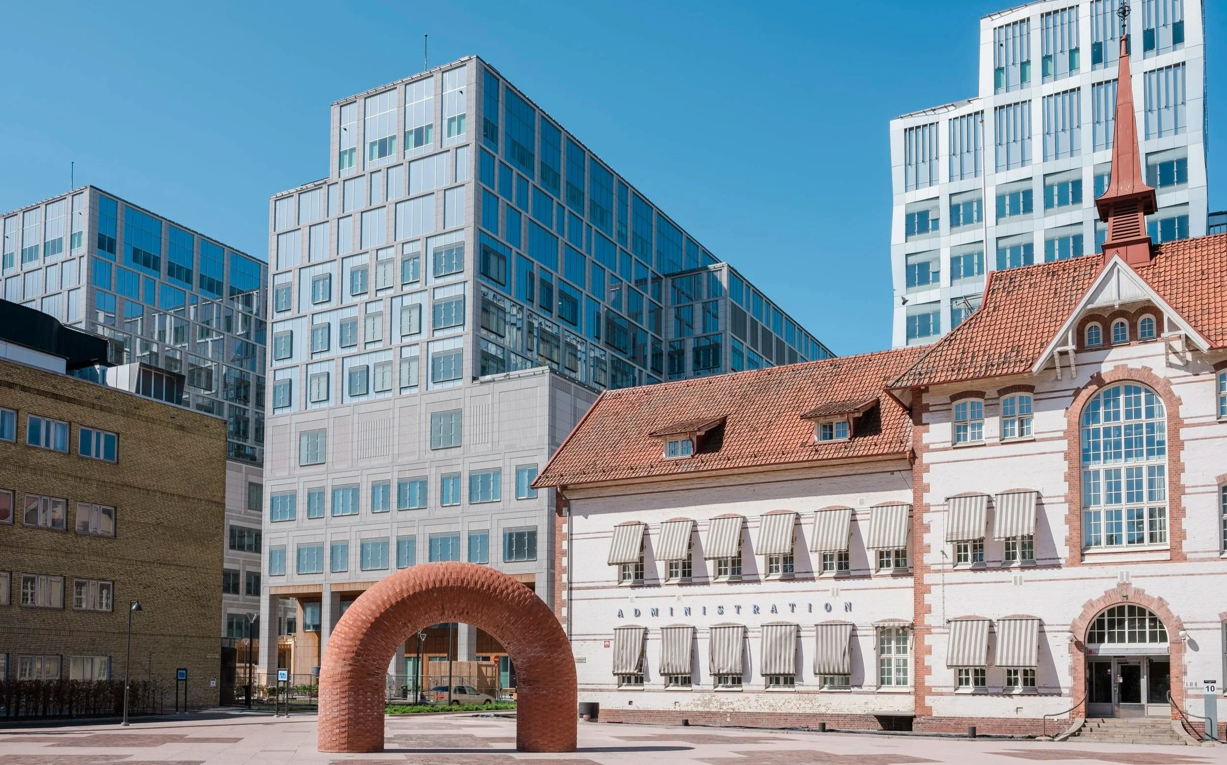 A cityscape with modern glass buildings and a historic building with a red-tiled roof, a steeple, and striped awnings, with a brick arch structure in the foreground and a blue sky overhead.