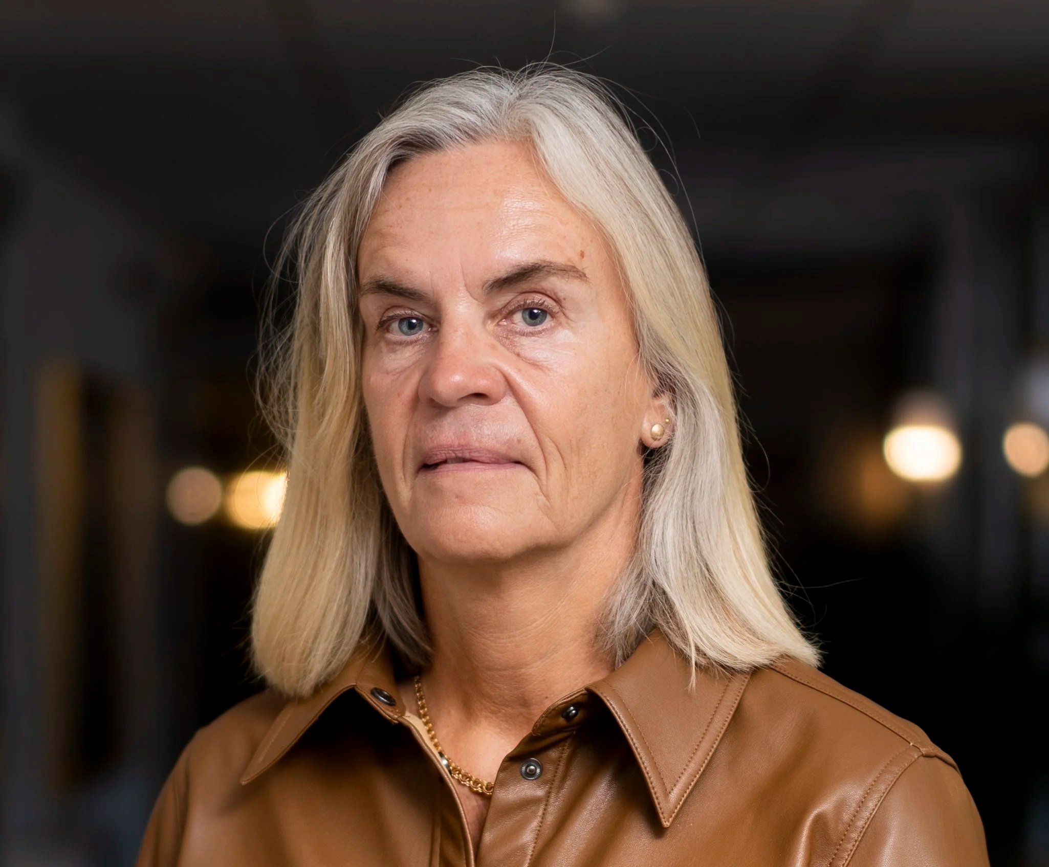 Close-up of a mature woman with silver hair, wearing a brown leather jacket, a gold necklace, and earrings, looking at the camera with a neutral expression, with a dark blurred background.