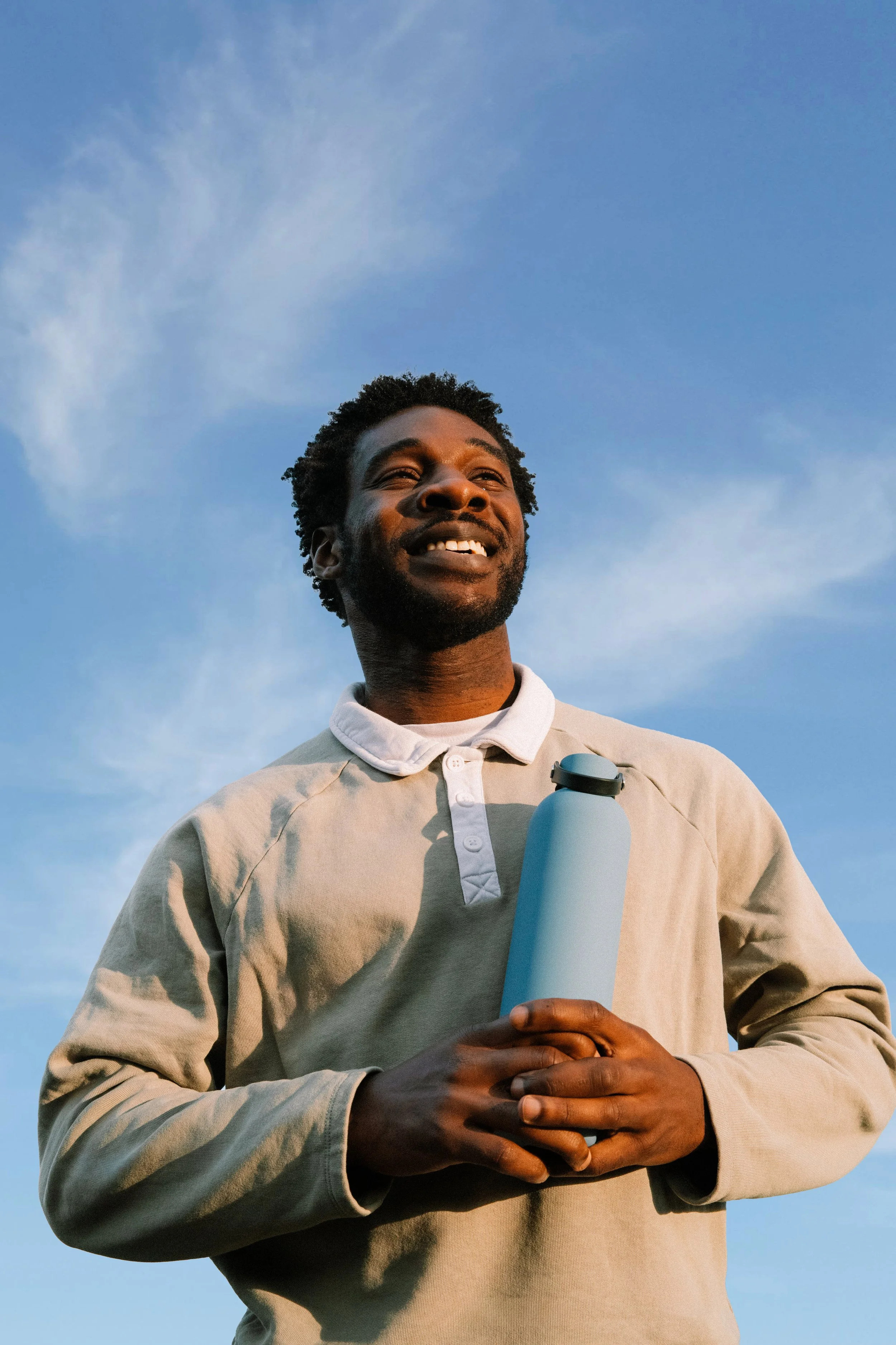 A man with short curly hair and a beard smiling outdoors, holding a water bottle, against a blue sky with some clouds.