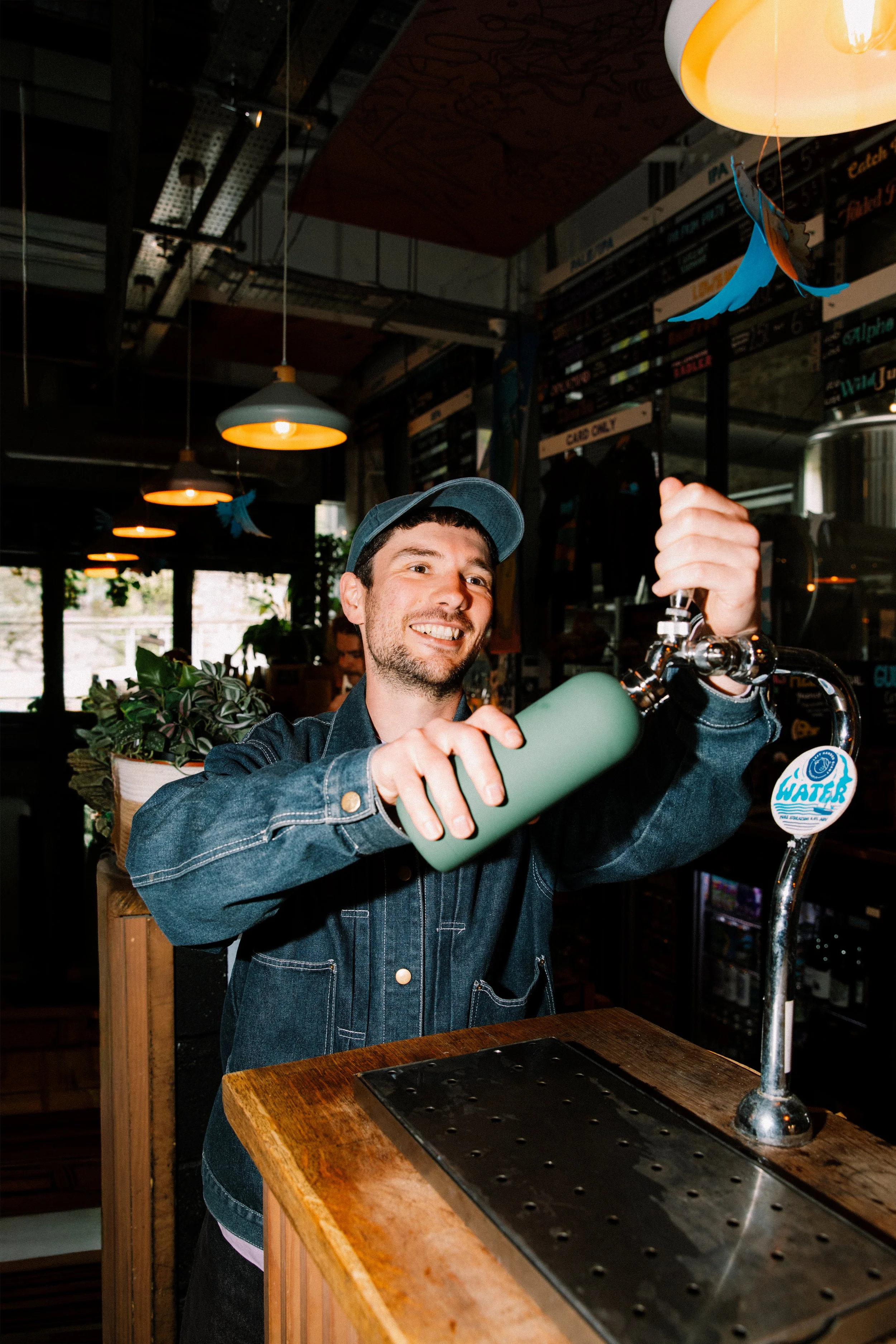 A man in a denim jacket and baseball cap is pouring water from a green water bottle into a glass at a bar or café counter. The setting has warm lighting, plants, and a menu board in the background.