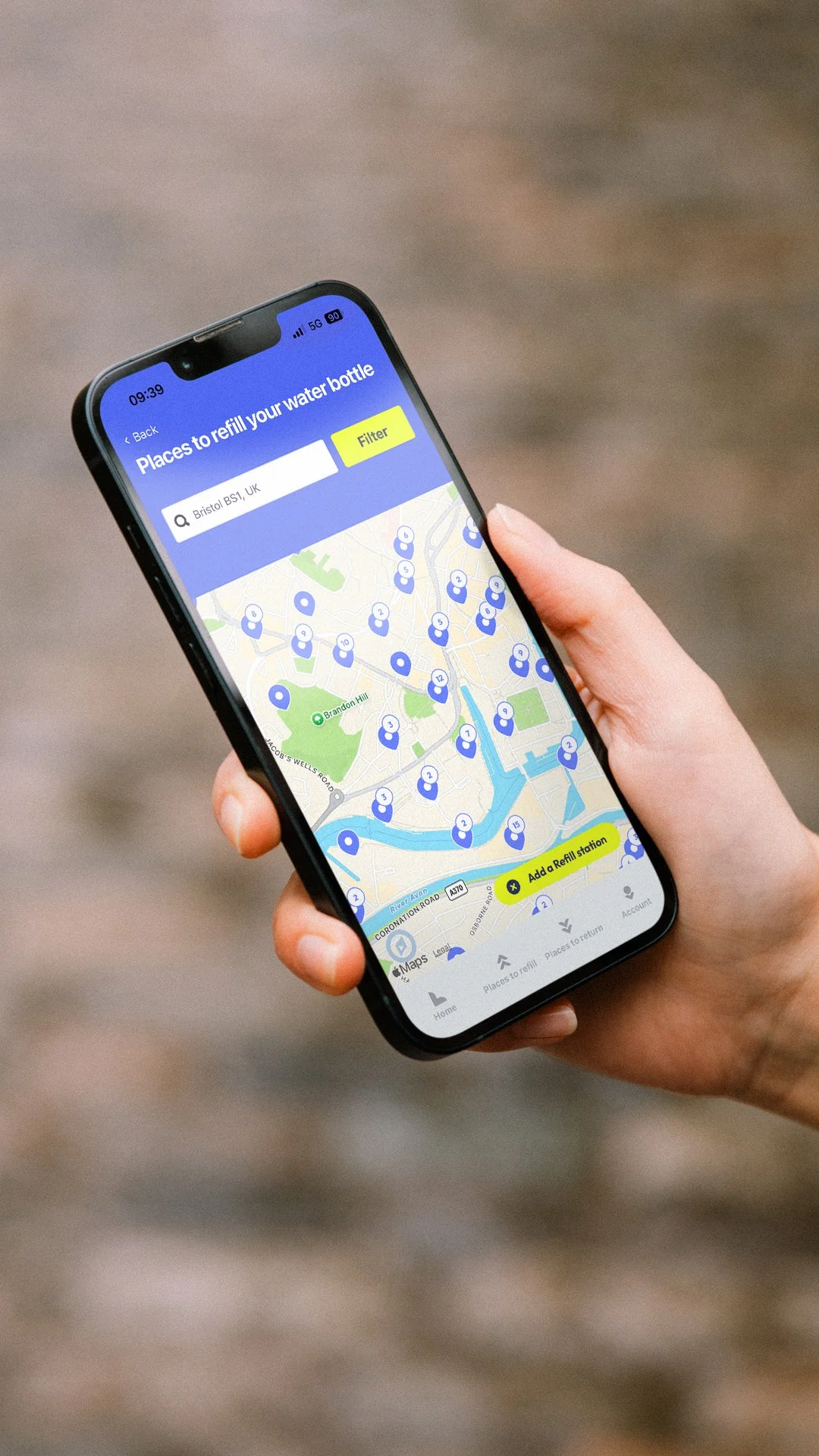 A person holding a smartphone displaying a map on an app for finding places to refill water bottles. The map shows numerous water refill station icons, with a search bar at the top and a yellow button at the bottom to add more refill stations.