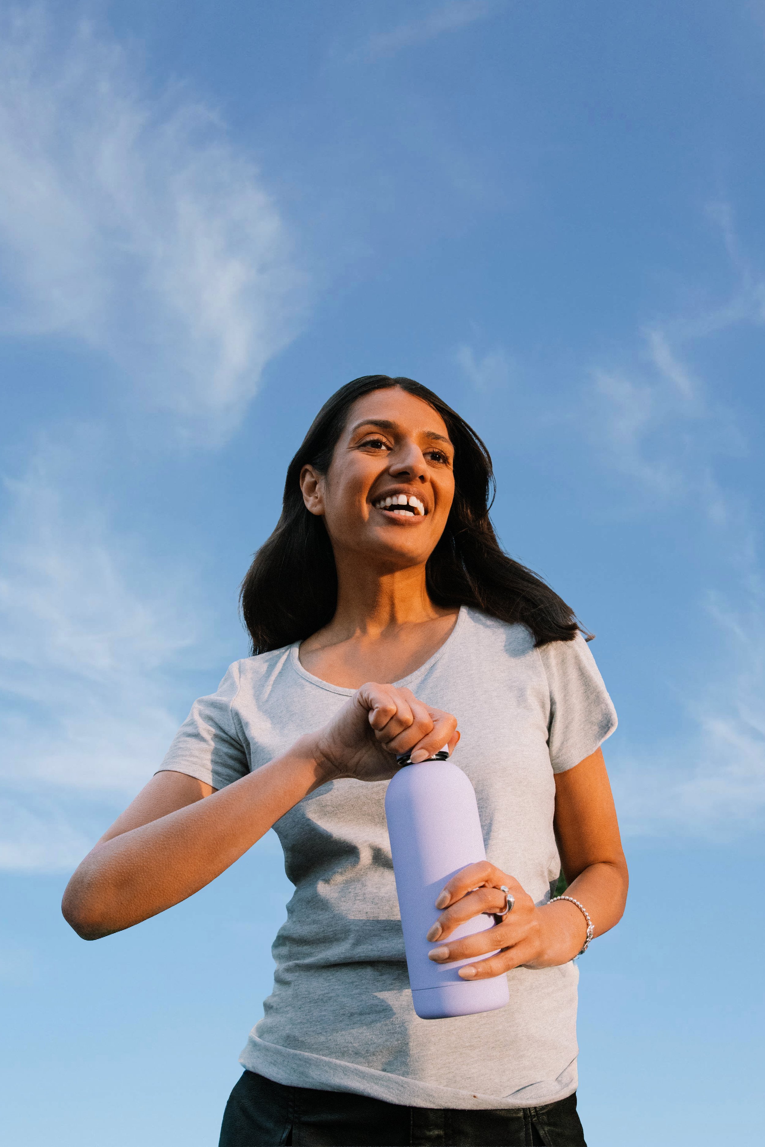 A woman with dark hair wearing a light gray t-shirt, holding a white water bottle, smiling, standing outside against a blue sky with light clouds.