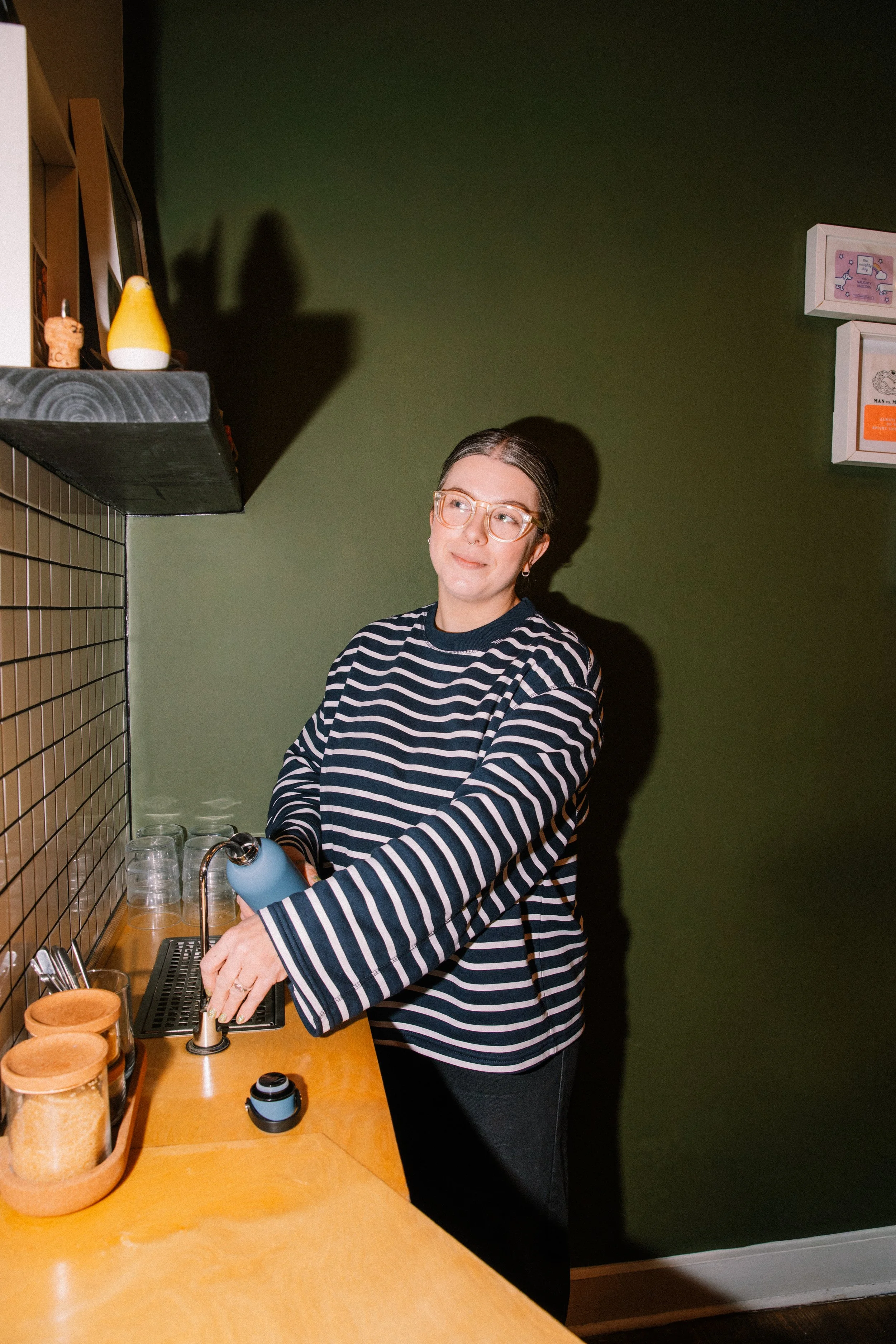 A woman wearing glasses and a black and white striped sweater is standing at a wooden counter, filling a cup with water from a faucet in a kitchen or coffee shop setting.