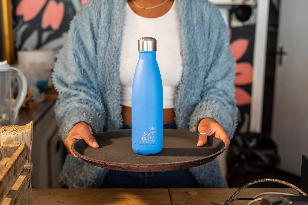 Person holding a black tray with a blue reusable water bottle on it in a kitchen setting.