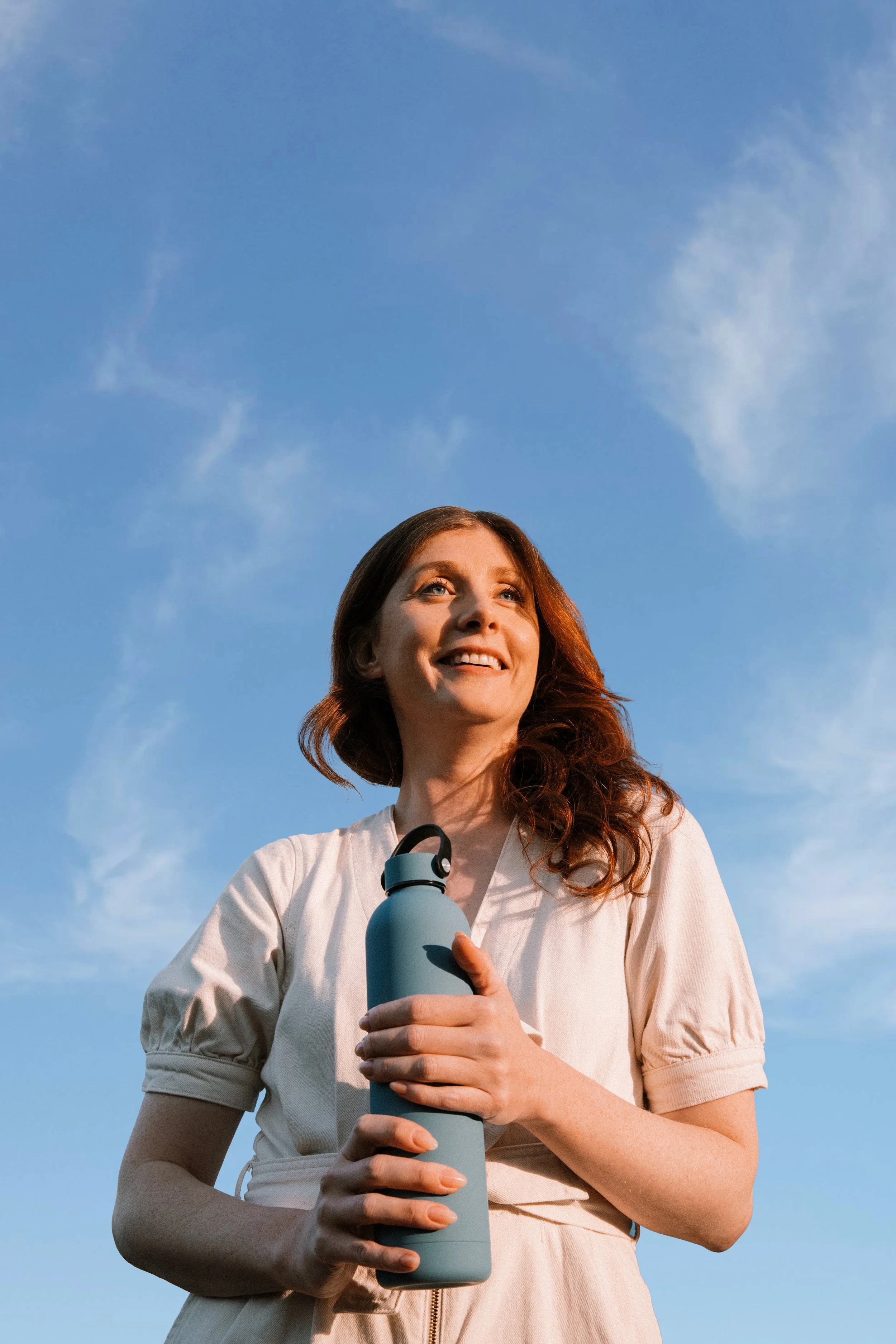 A woman with red hair smiling outdoors, holding a light blue water bottle, against a blue sky with a few clouds.