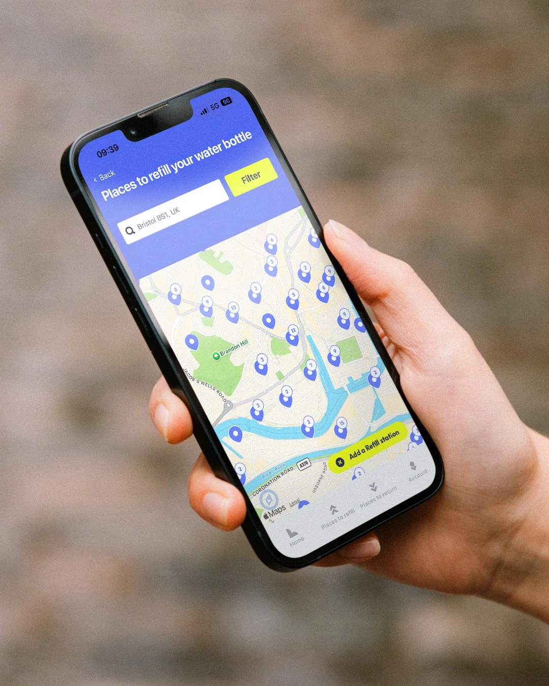A person holding a smartphone displaying a map with blue water refill station icons, search bar, filter button, and 'Add a Refill station' button for finding water refill stations in Bristol, UK.
