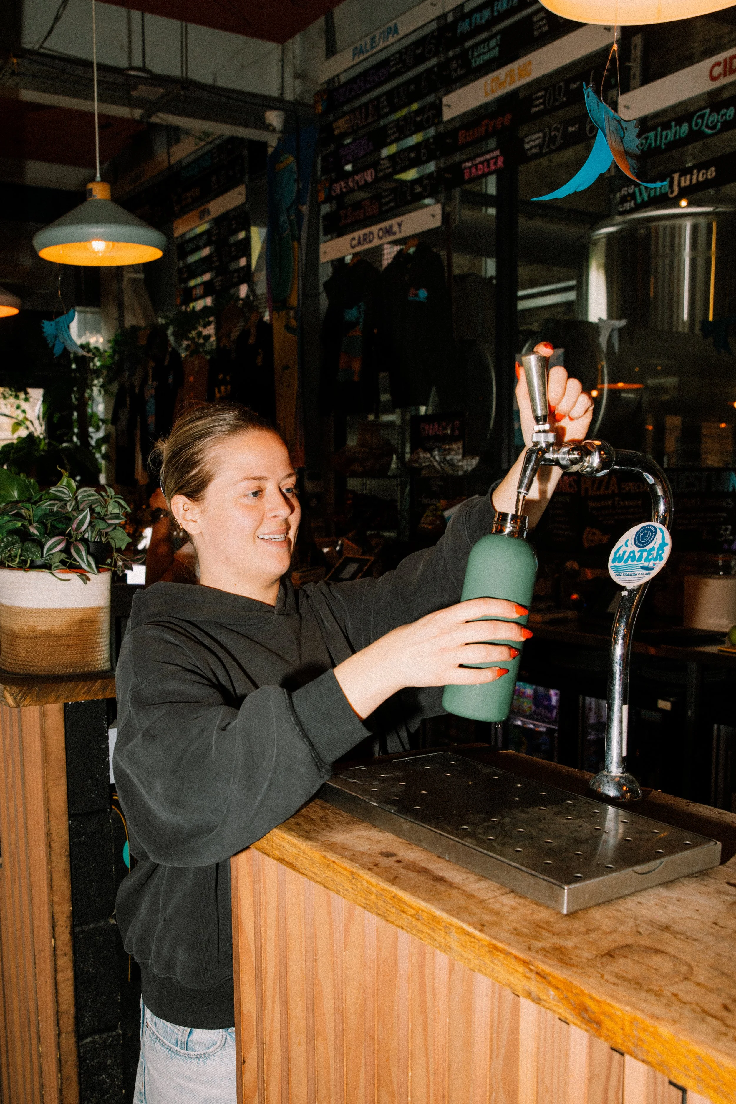 A woman with light brown hair tied back, wearing a black hoodie, is filling a green reusable water bottle at a bar tap in a casual indoor setting. The environment features a wooden counter, plants, and a menu board with various items and prices in the background.
