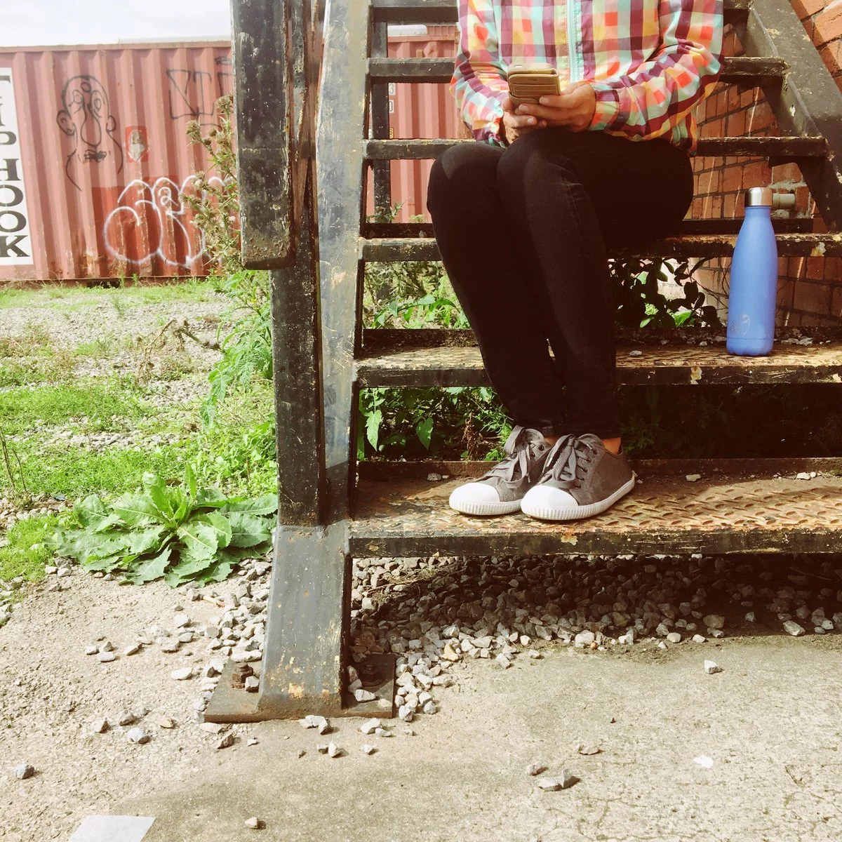 Person sitting on outdoor metal staircase using a smartphone, wearing a multicolored checkered jacket, black pants, and gray sneakers, with a blue water bottle beside them, and graffiti on a red container in the background.